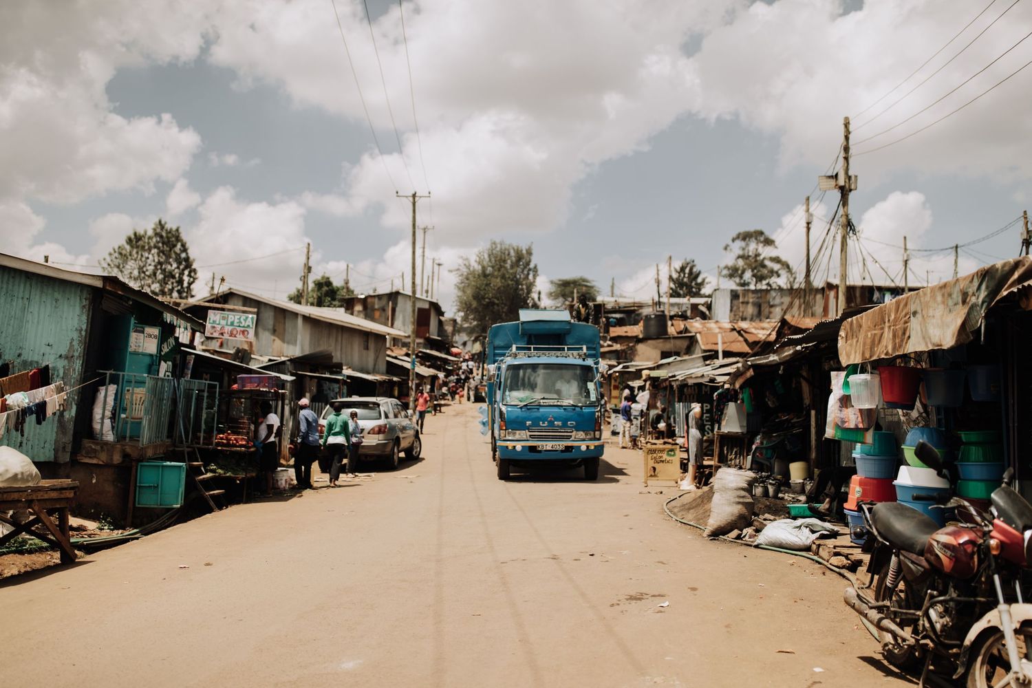 A dusty street scene in Haiti shows local shops and buildings with blue trucks parked along an unpaved road.