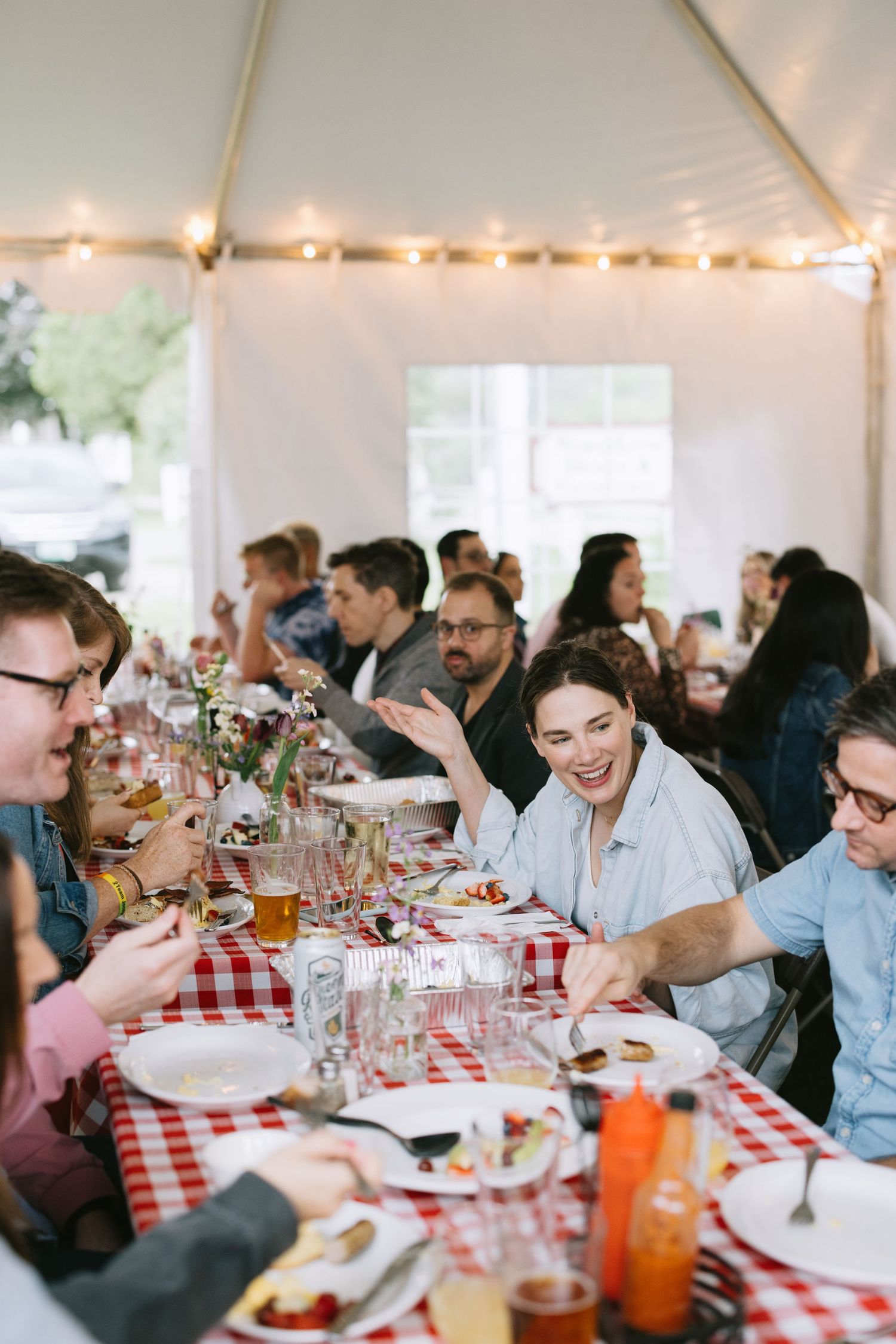 Guests enjoy a casual dinner under string lights at red and white checkered tables during a festive gathering.