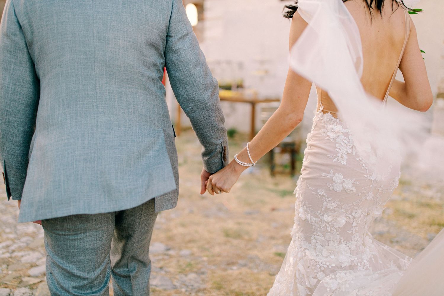 A couple in wedding attire hold hands while walking together, with the groom in a gray suit and bride in a white dress.