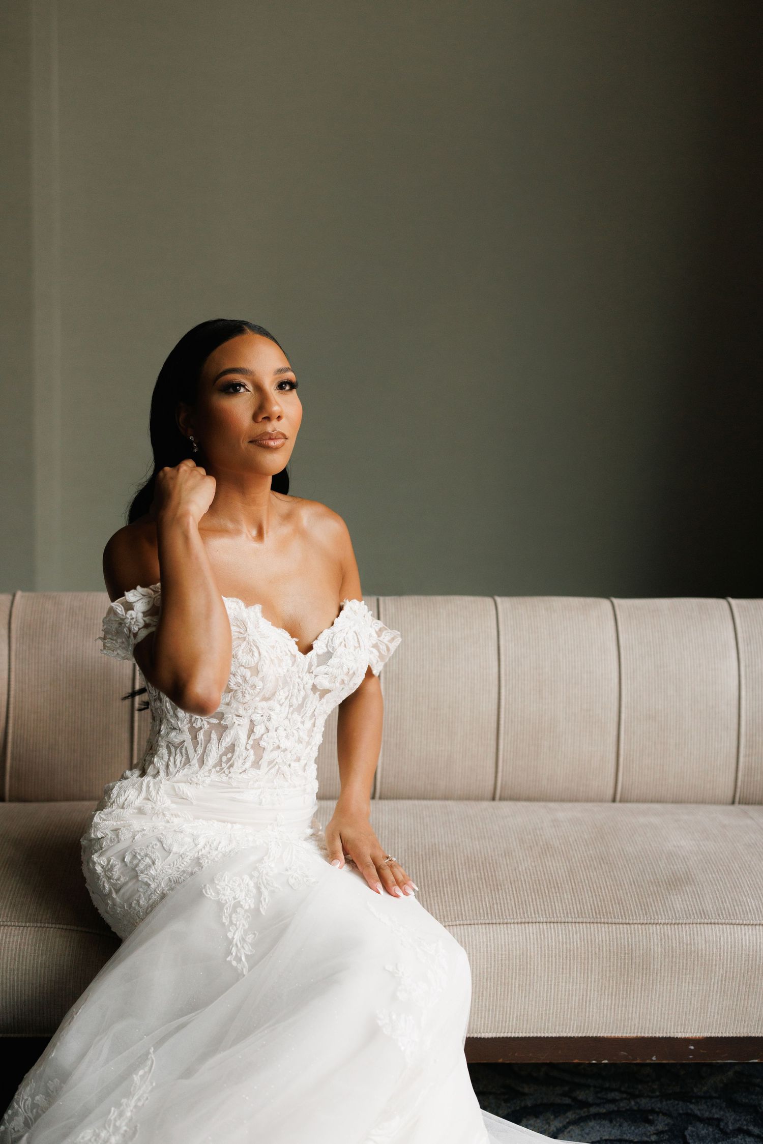 A stunning bride in an off-shoulder lace wedding gown with floral details sits elegantly on a beige couch.