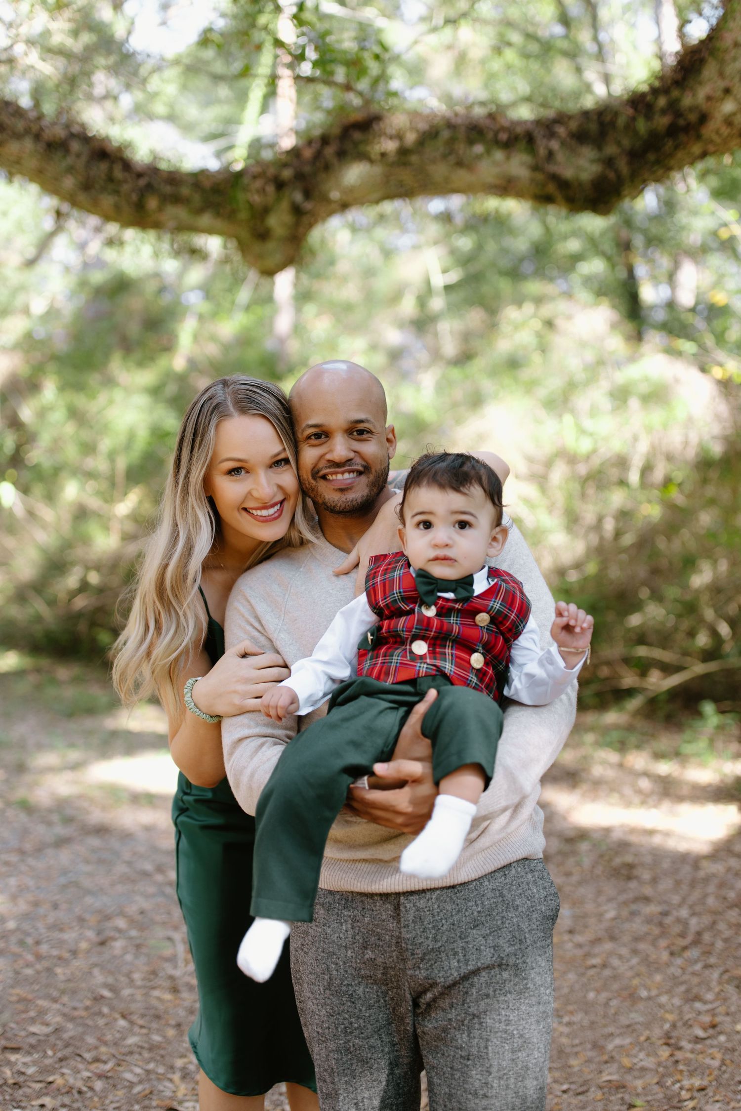 A family poses together outdoors under a large tree branch in matching neutral-toned outfits on a sunny day.