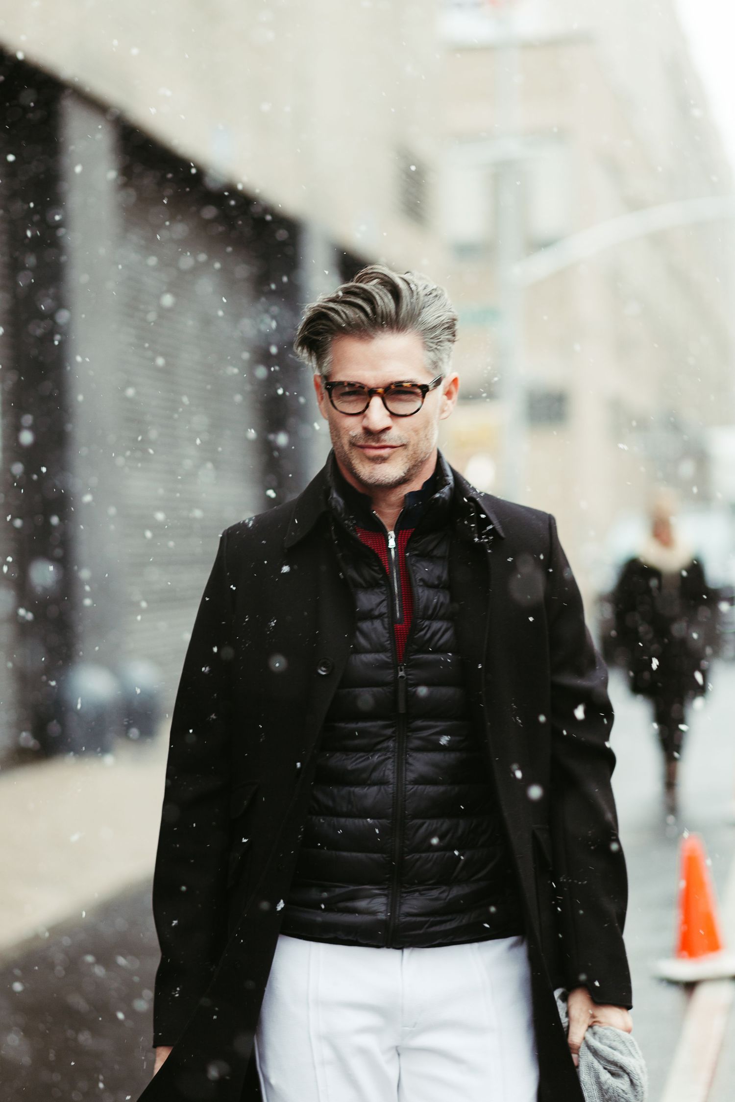 Stylish winter street fashion featuring a layered look with black puffer vest, wool coat and glasses during snowfall.