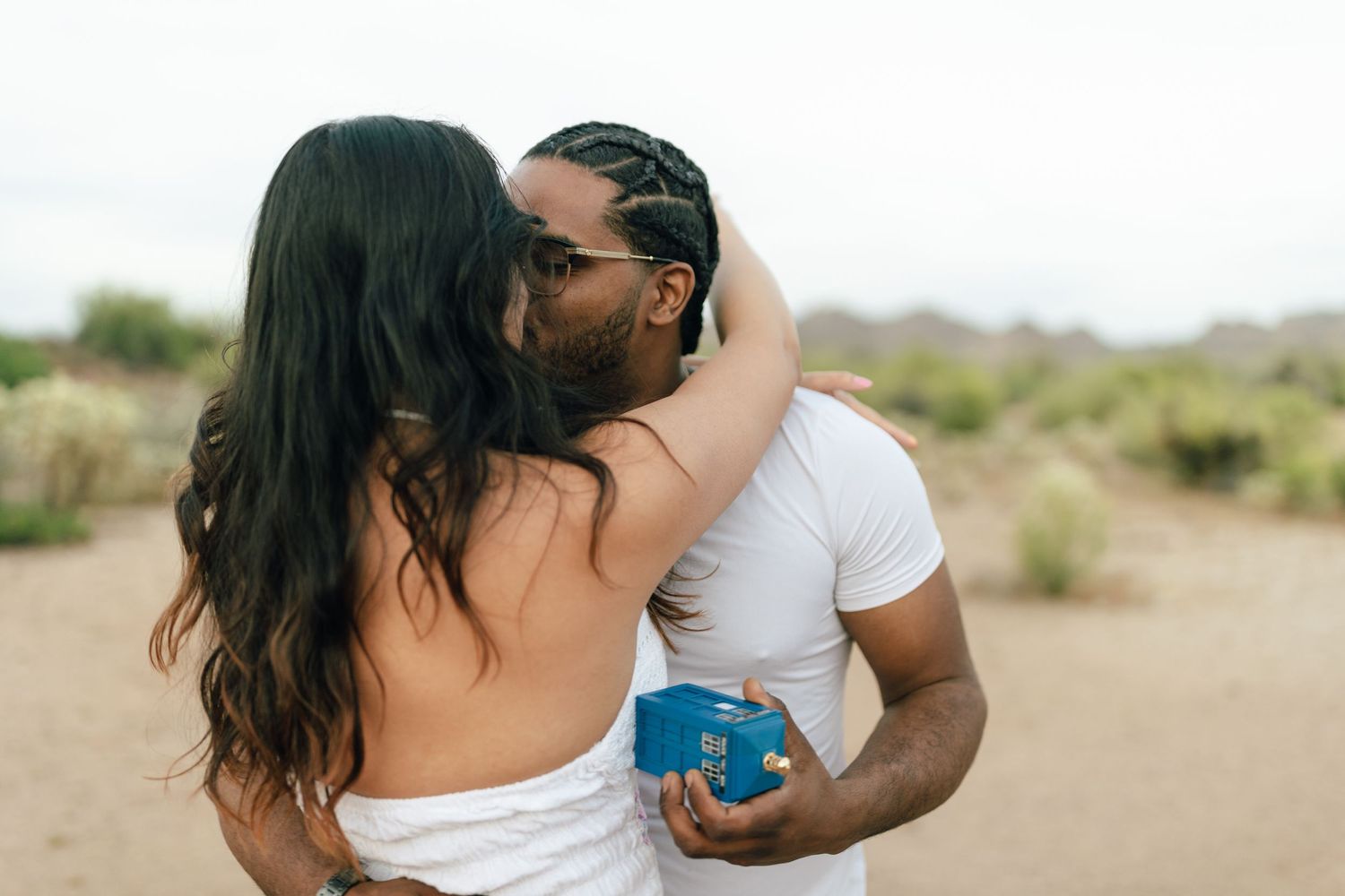 A romantic couple embraces in an outdoor desert setting with natural brush in the background.