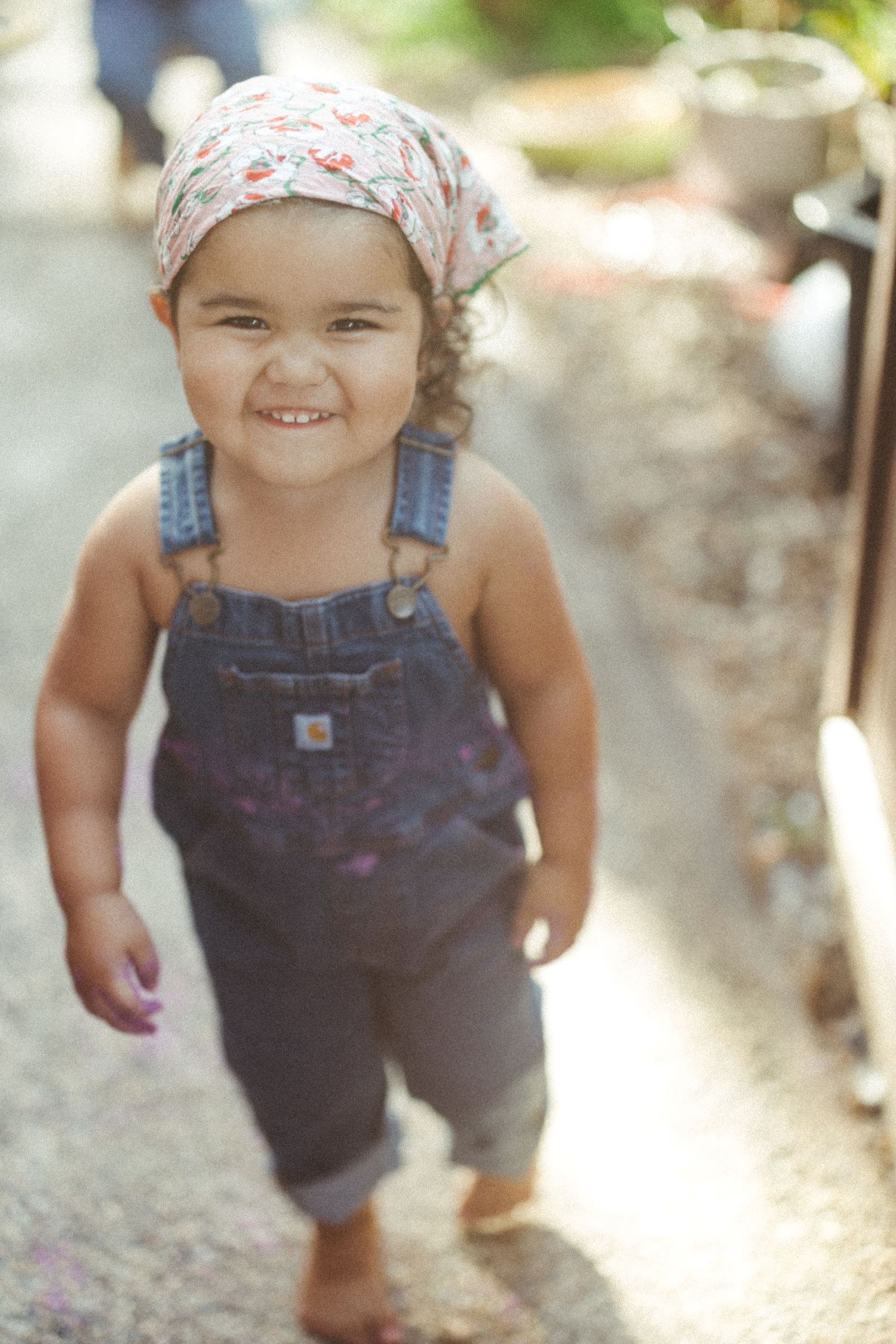 A small child wearing a bandana and denim overalls smiles brightly while walking on a sunny outdoor path.