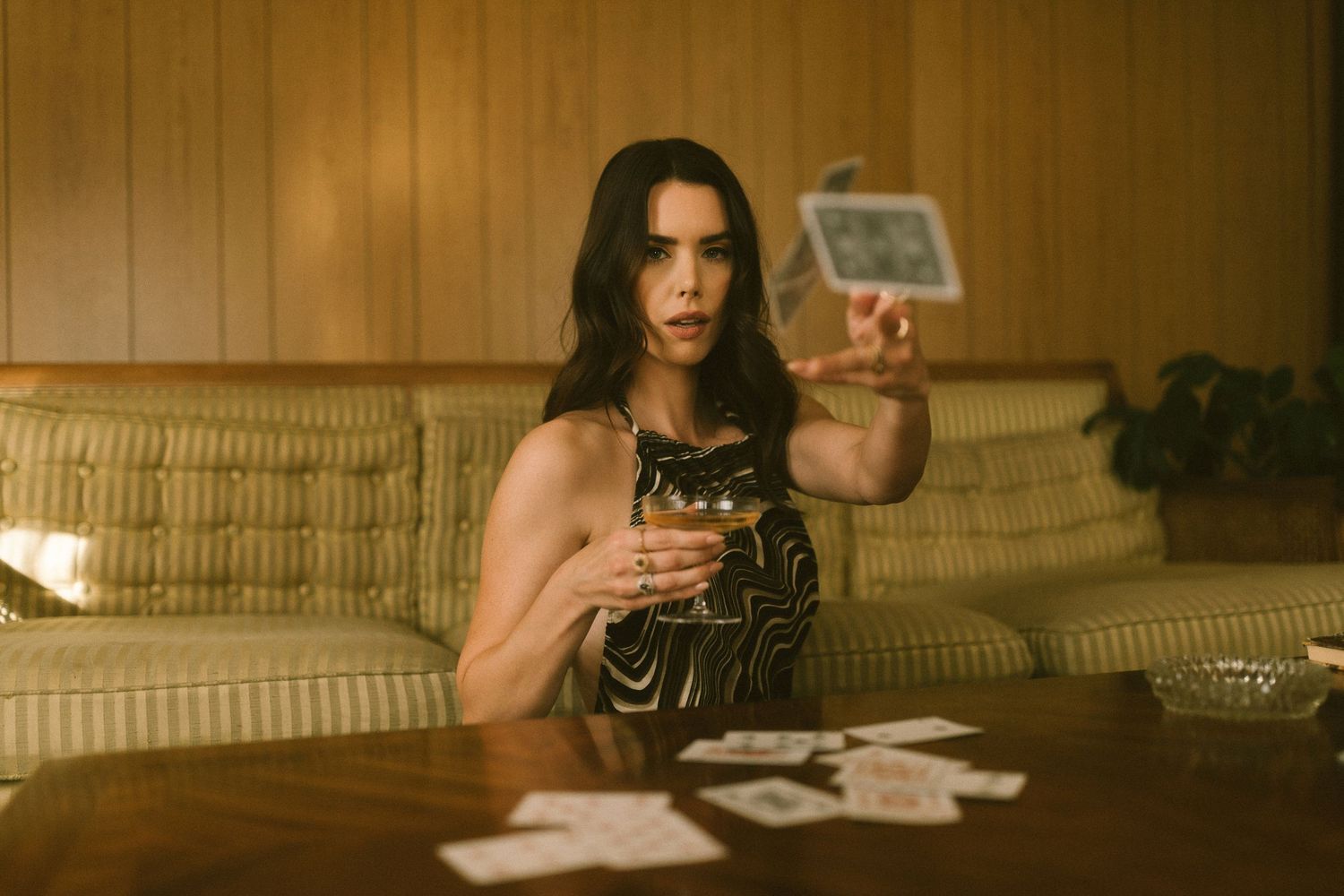 Someone holds a handgun while sitting on a vintage couch in a dimly lit room with playing cards on the table.