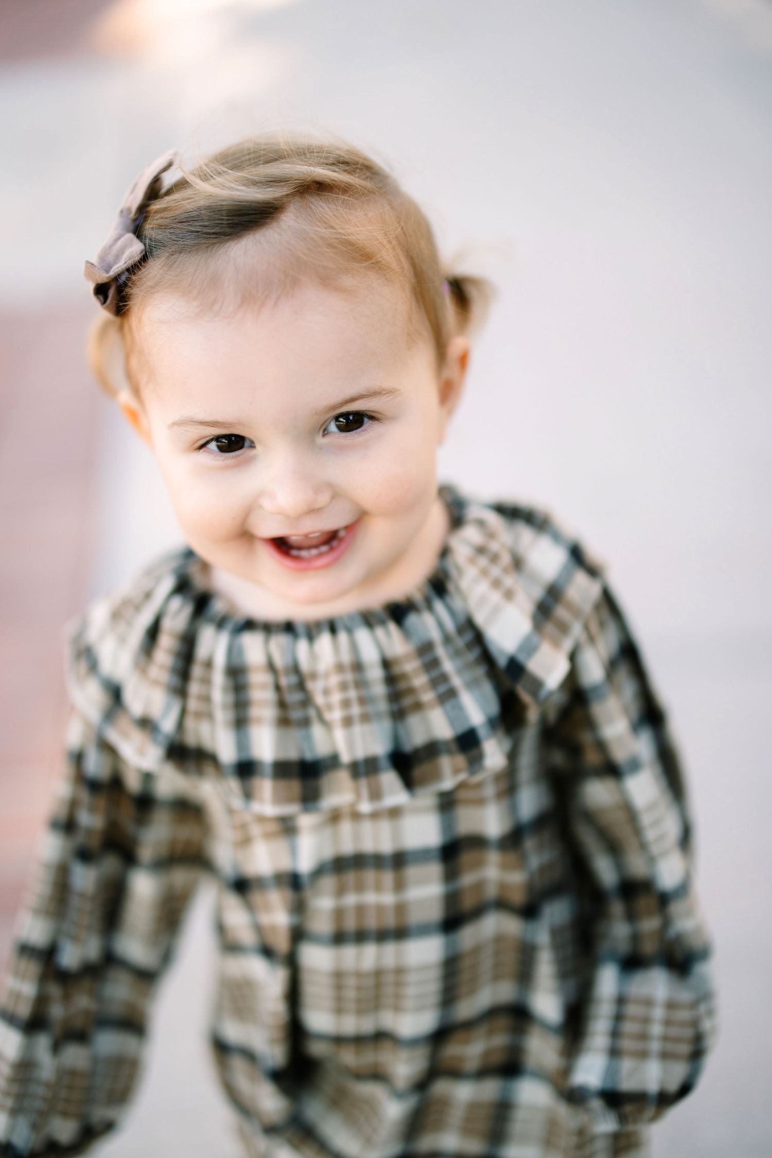 Joyful little toddler wearing a plaid dress with a small hair clip smiles brightly at the camera.