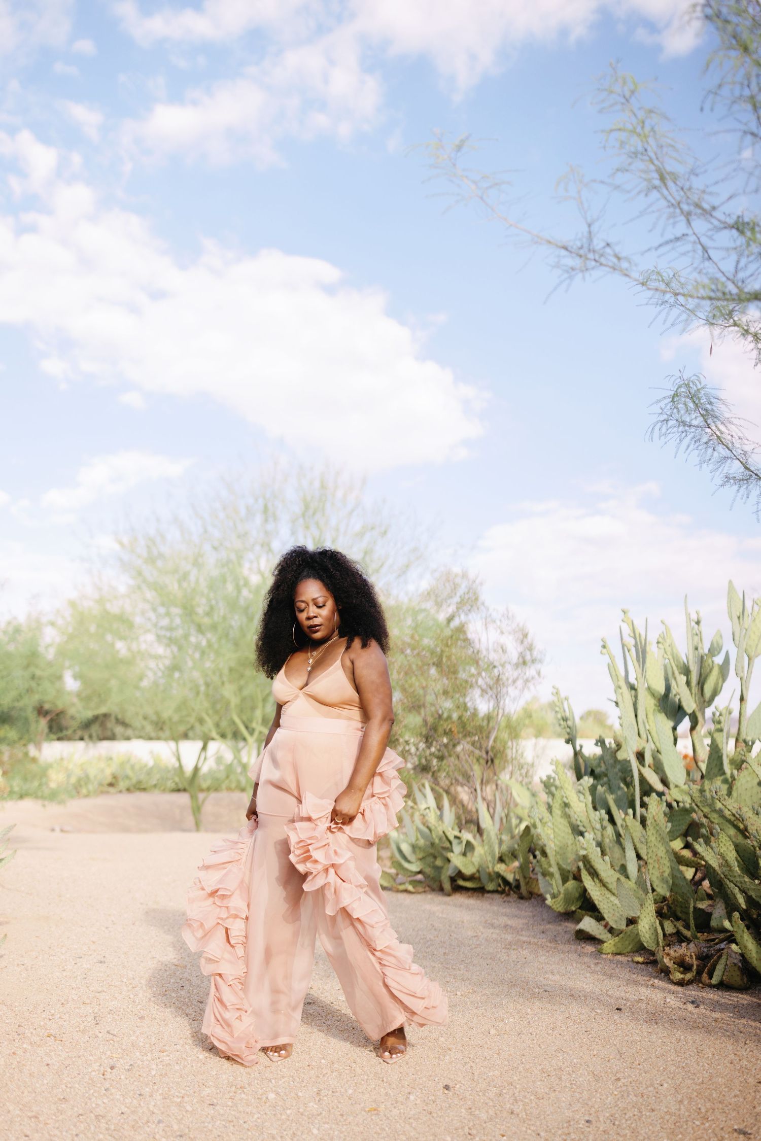 A person in a flowing peach dress dances gracefully in a desert garden setting with cacti and succulents under blue skies.