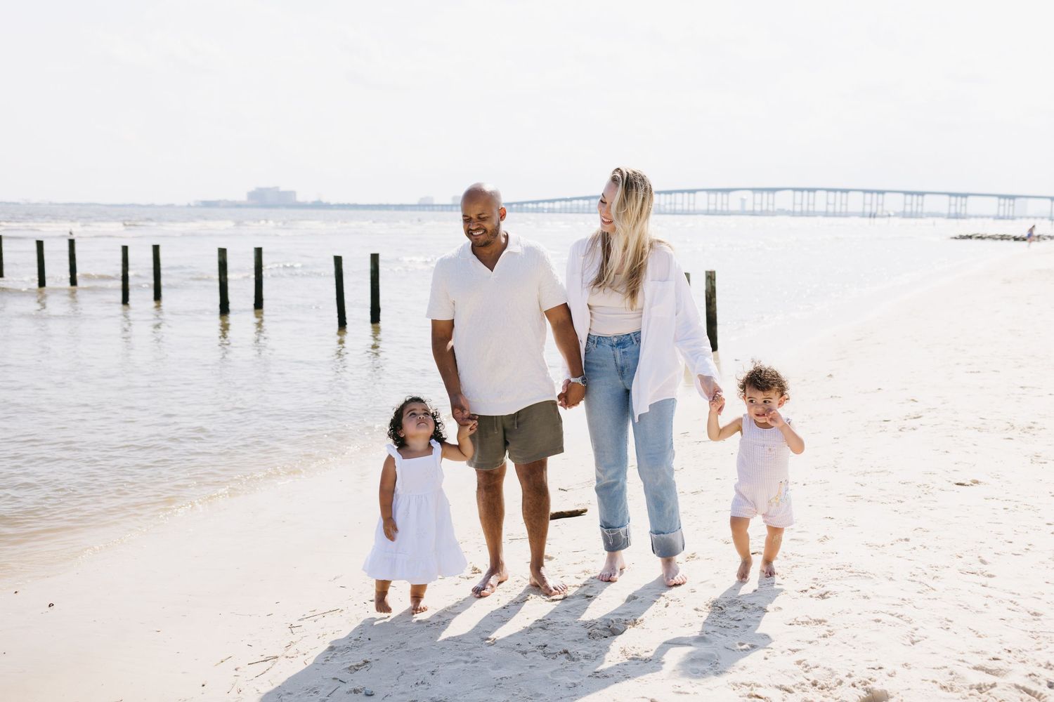 Family walking together on white sandy beach during sunset while wearing casual summer clothes and casting long shadows.