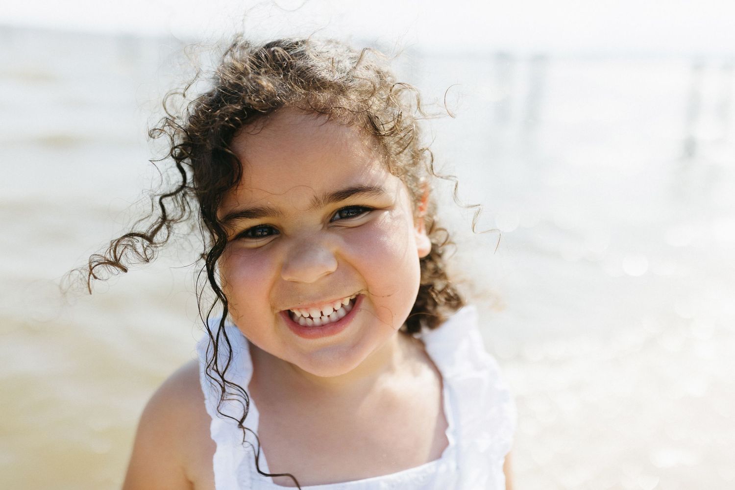 Young child with curly dark hair wearing white smiles brightly in natural outdoor lighting.