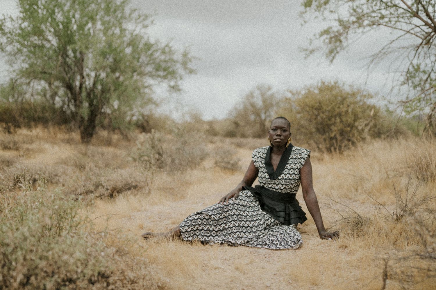 A figure in a patterned dress sits thoughtfully in a desert landscape with sparse vegetation and muted tones.