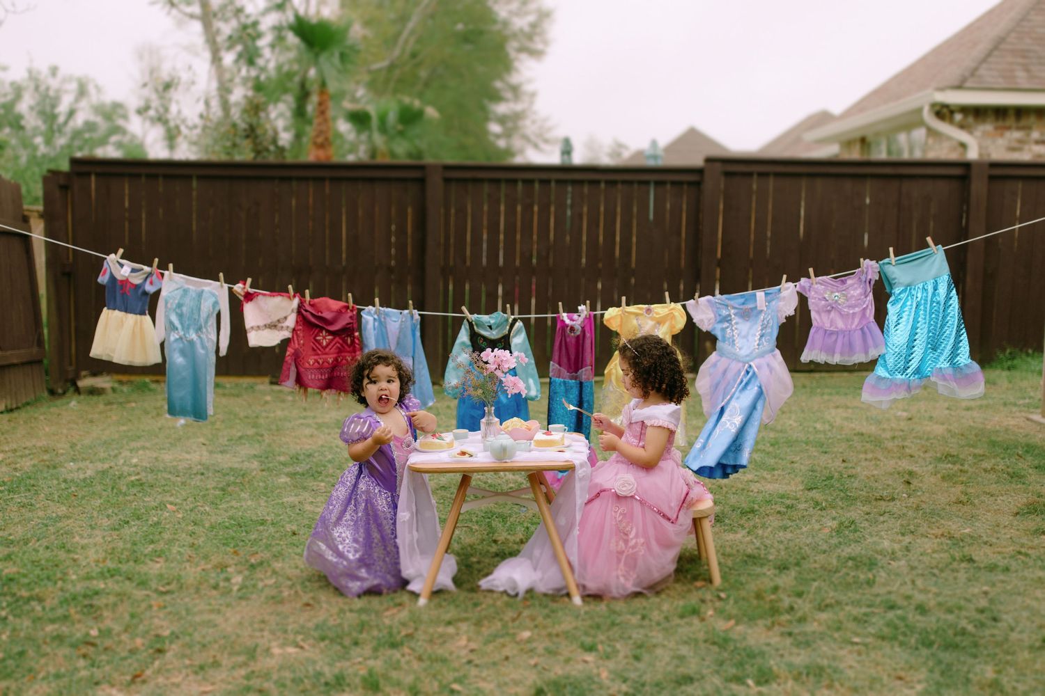 Colorful children's dresses and clothing hang from an outdoor clothesline against a wooden fence in a backyard.