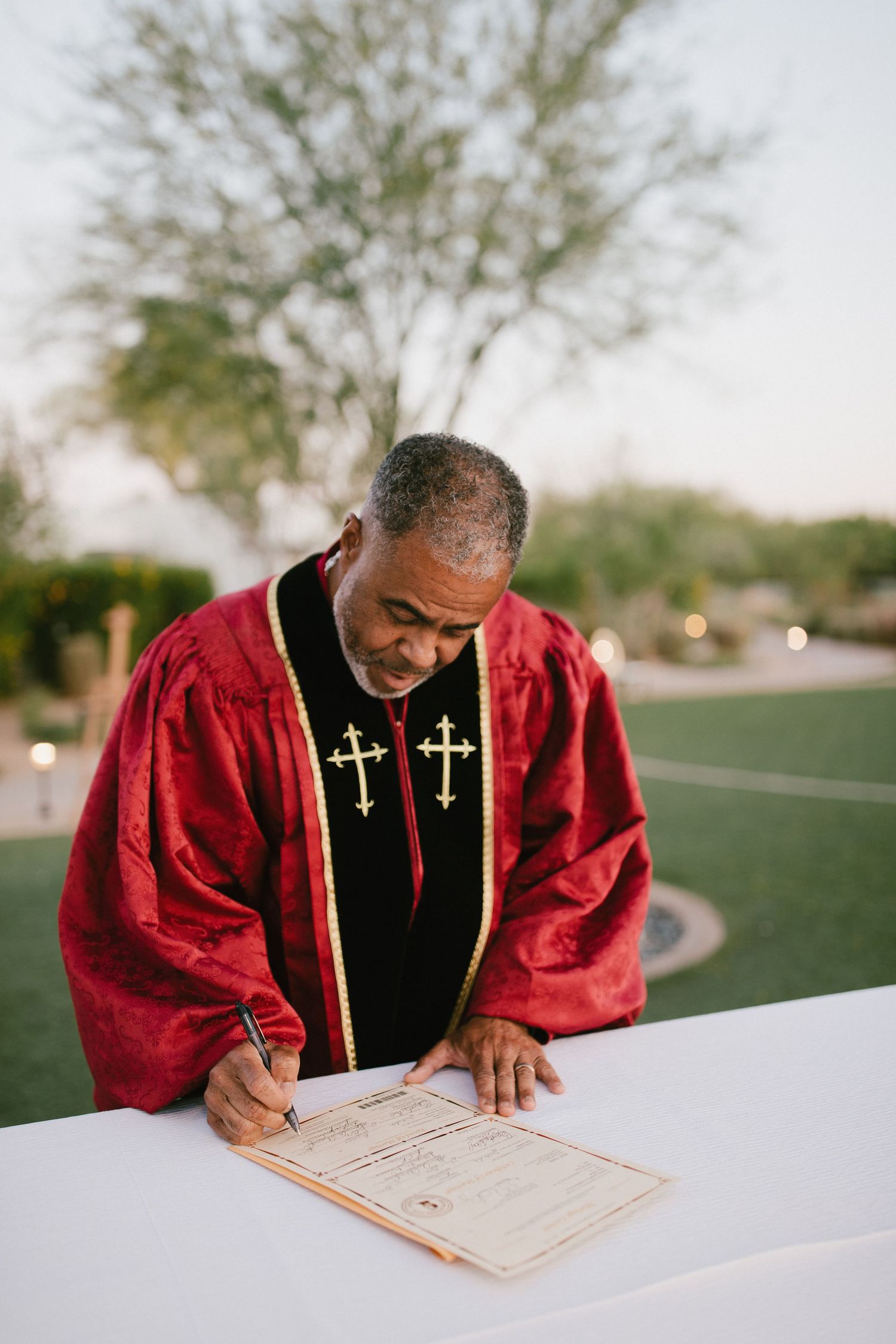 Minister in red and black robe with cross embellishments signs document at white table outdoors.