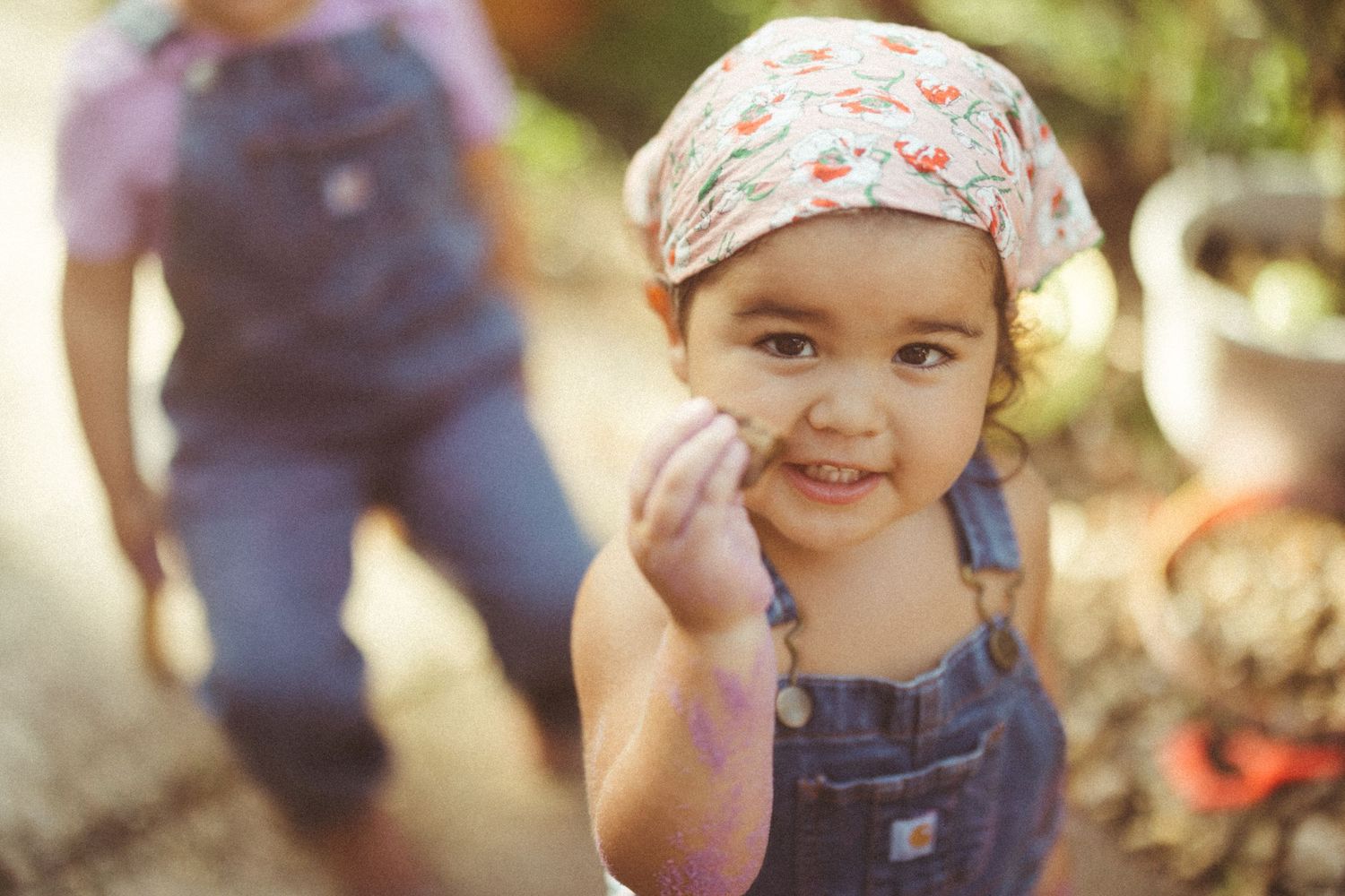 A young child in overalls and a floral headscarf smiles sweetly outdoors in natural lighting.