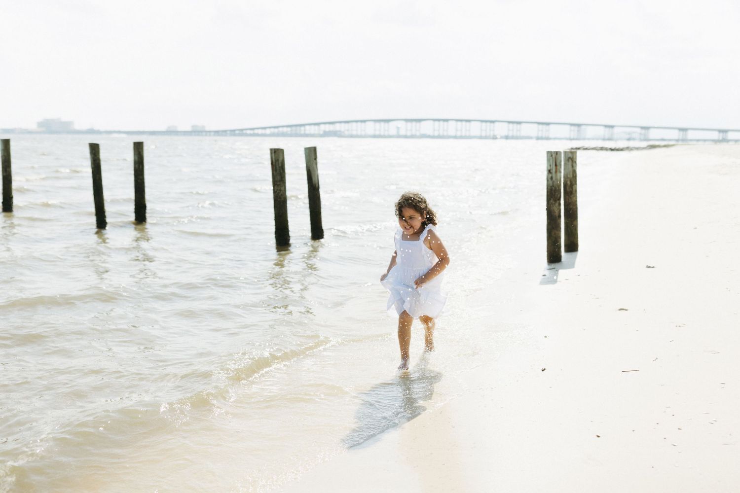 A person in a white dress walks along a misty beach with wooden pier posts in the background.