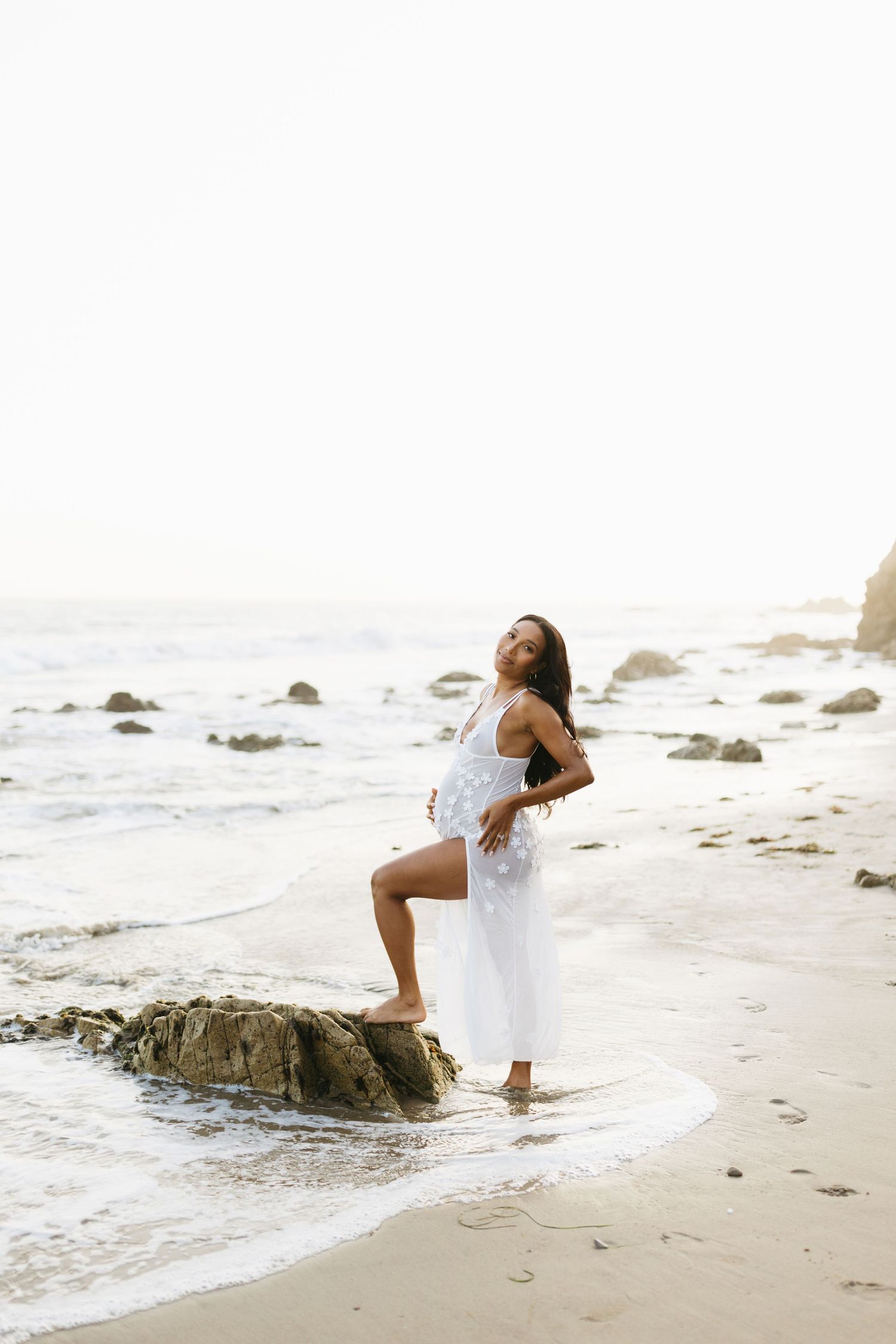 Woman in white dress poses gracefully on beach rocks during sunset photoshoot with waves crashing in background.