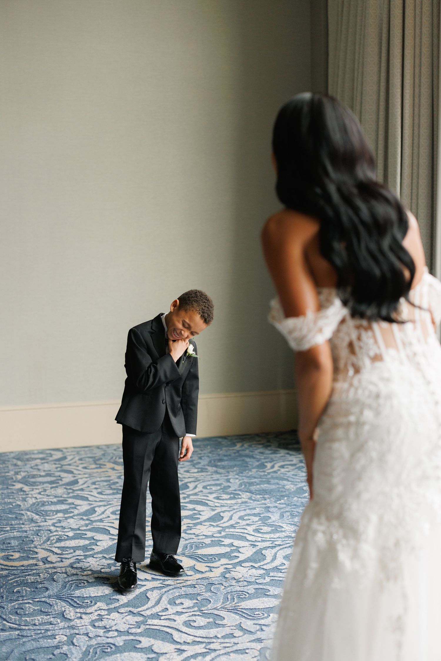 A young boy in a suit gets emotional upon seeing someone in a white lace wedding dress standing nearby.