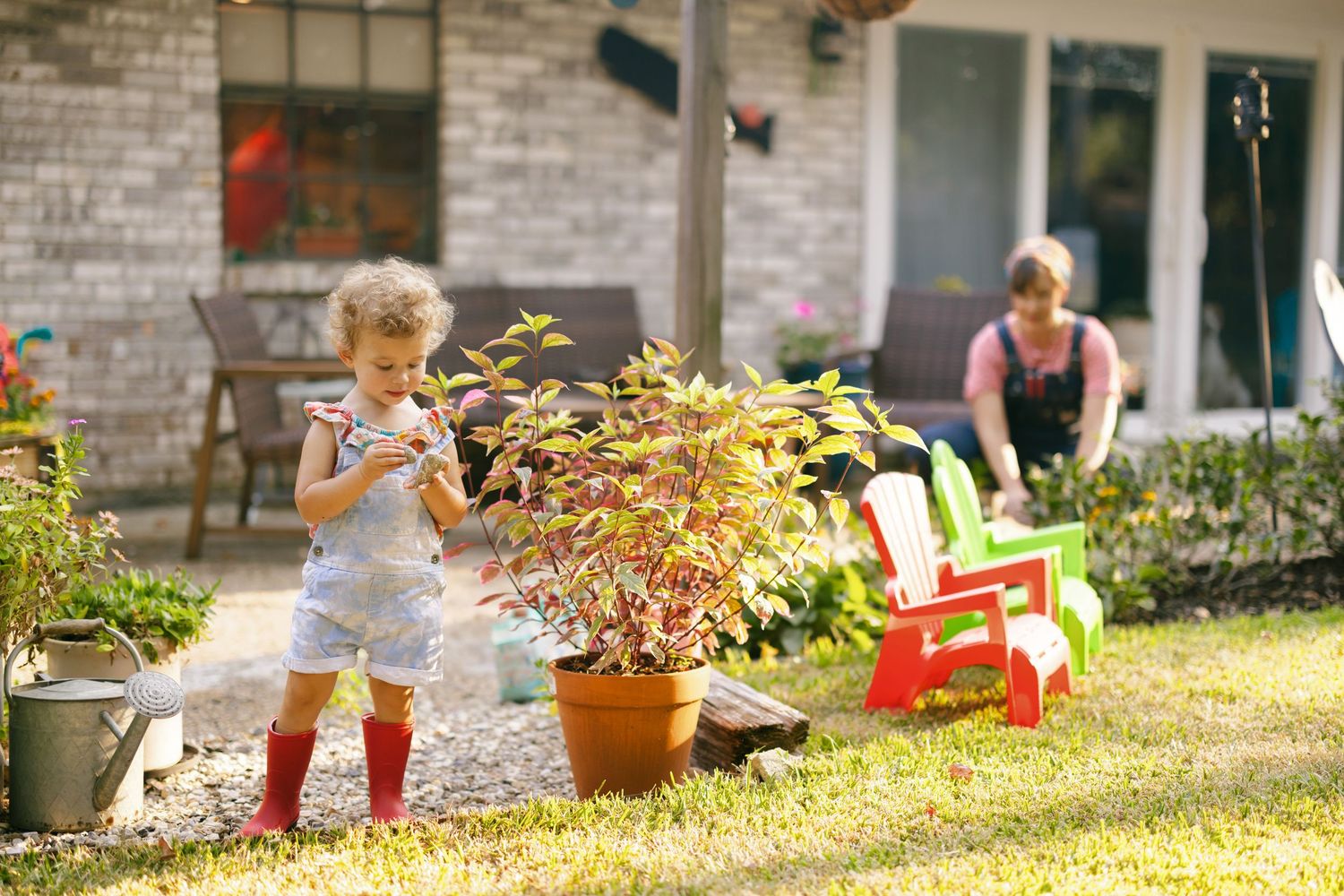 A child in red rain boots playing with water in a sunny garden with red Adirondack chairs and potted plants.