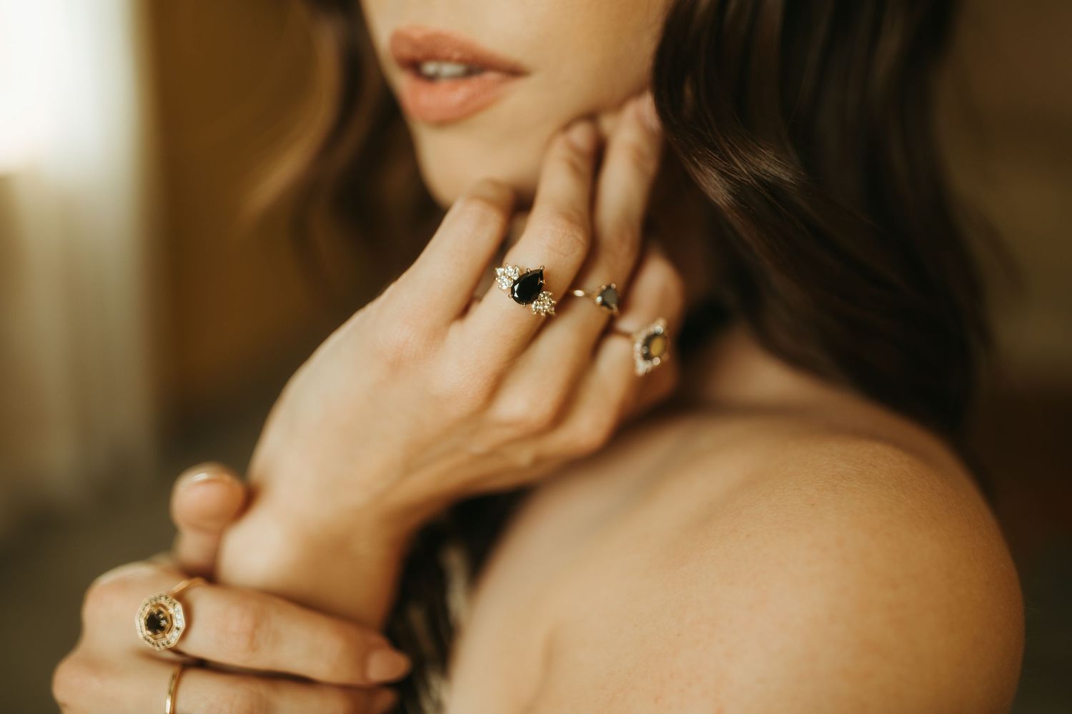 Close up beauty shot of lips and hands wearing intricate gold rings and dark nail polish in warm lighting.