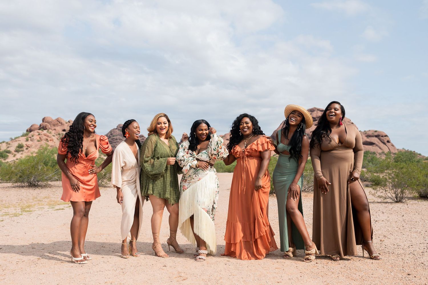 A group of women in casual dresses stand together laughing against a desert mountain backdrop on a sunny day.