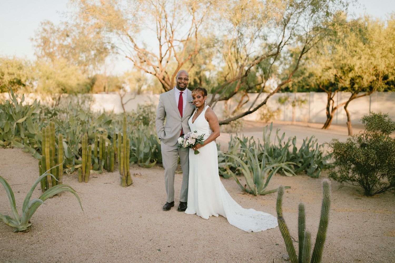 A couple poses together in a desert garden setting surrounded by tall saguaro cacti at golden hour.