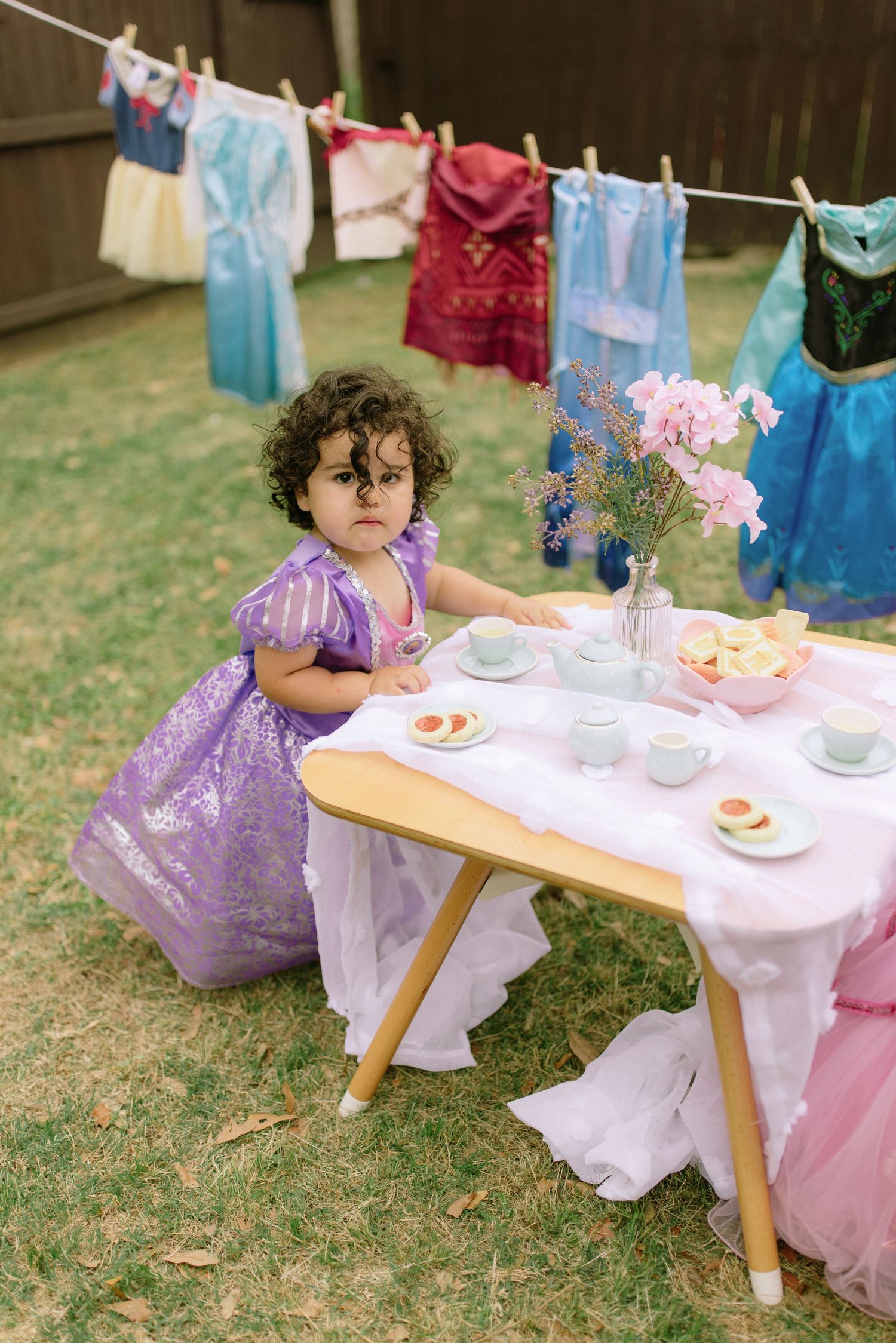 Tea party setup in backyard with fancy dress and laundry line of colorful princess dresses hanging in background.