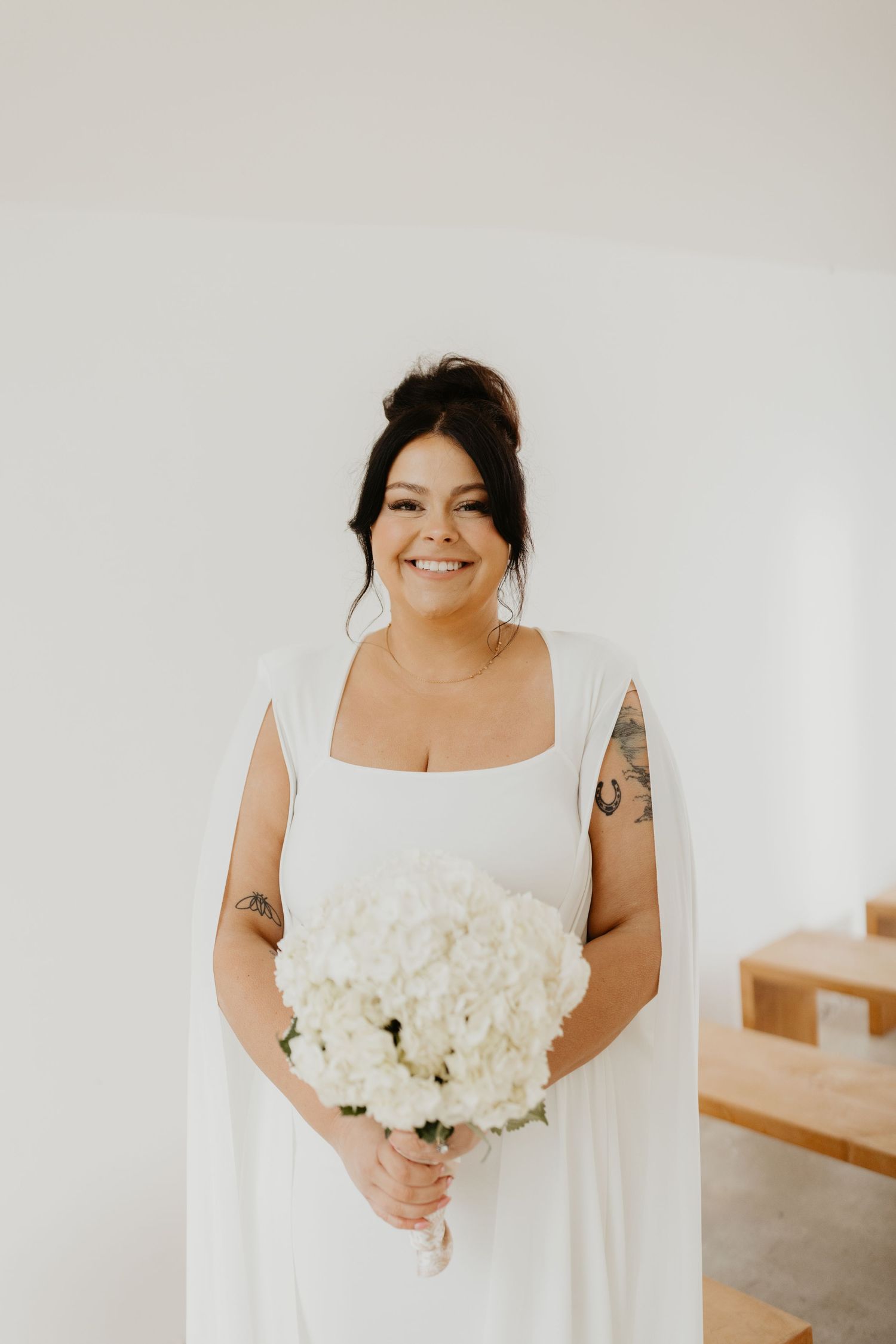 Bride in elegant white square-neck dress holds beautiful bouquet of white hydrangeas against neutral backdrop.