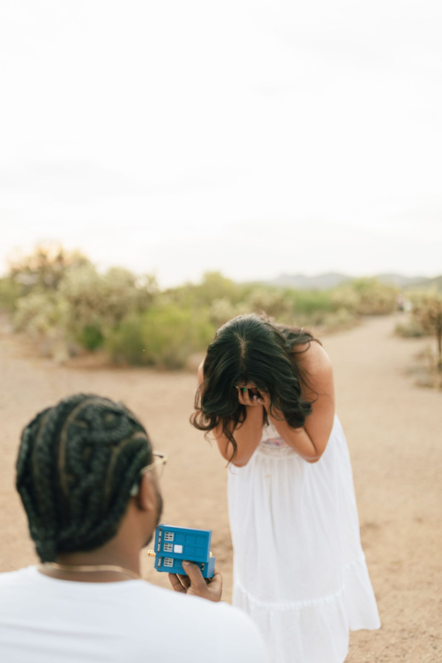 A marriage proposal moment captured on a desert trail during sunset with one person presenting a small blue box.
