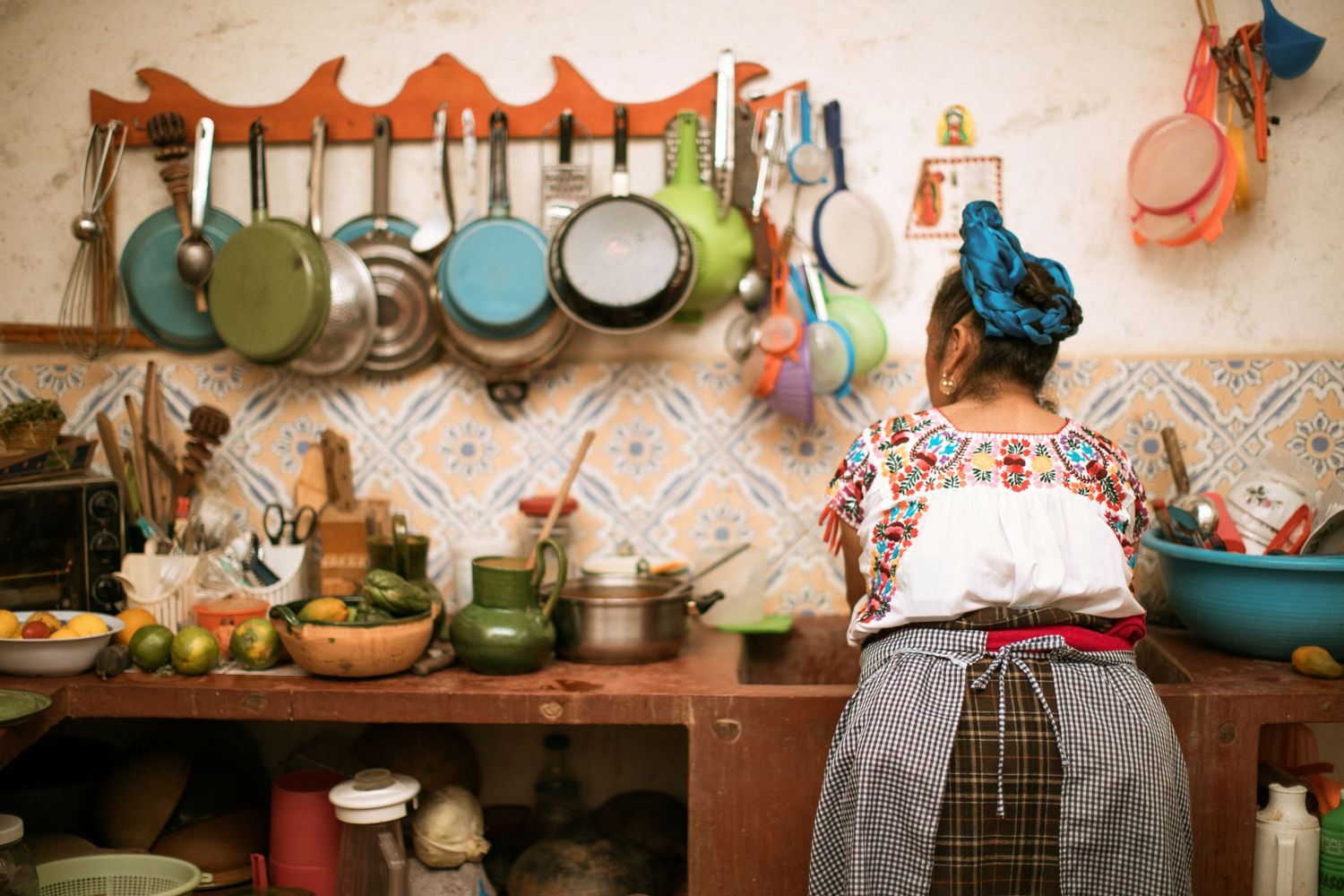 A vintage kitchen interior with copper pots hanging on the wall and a figure in floral print working at a rustic wooden counter.