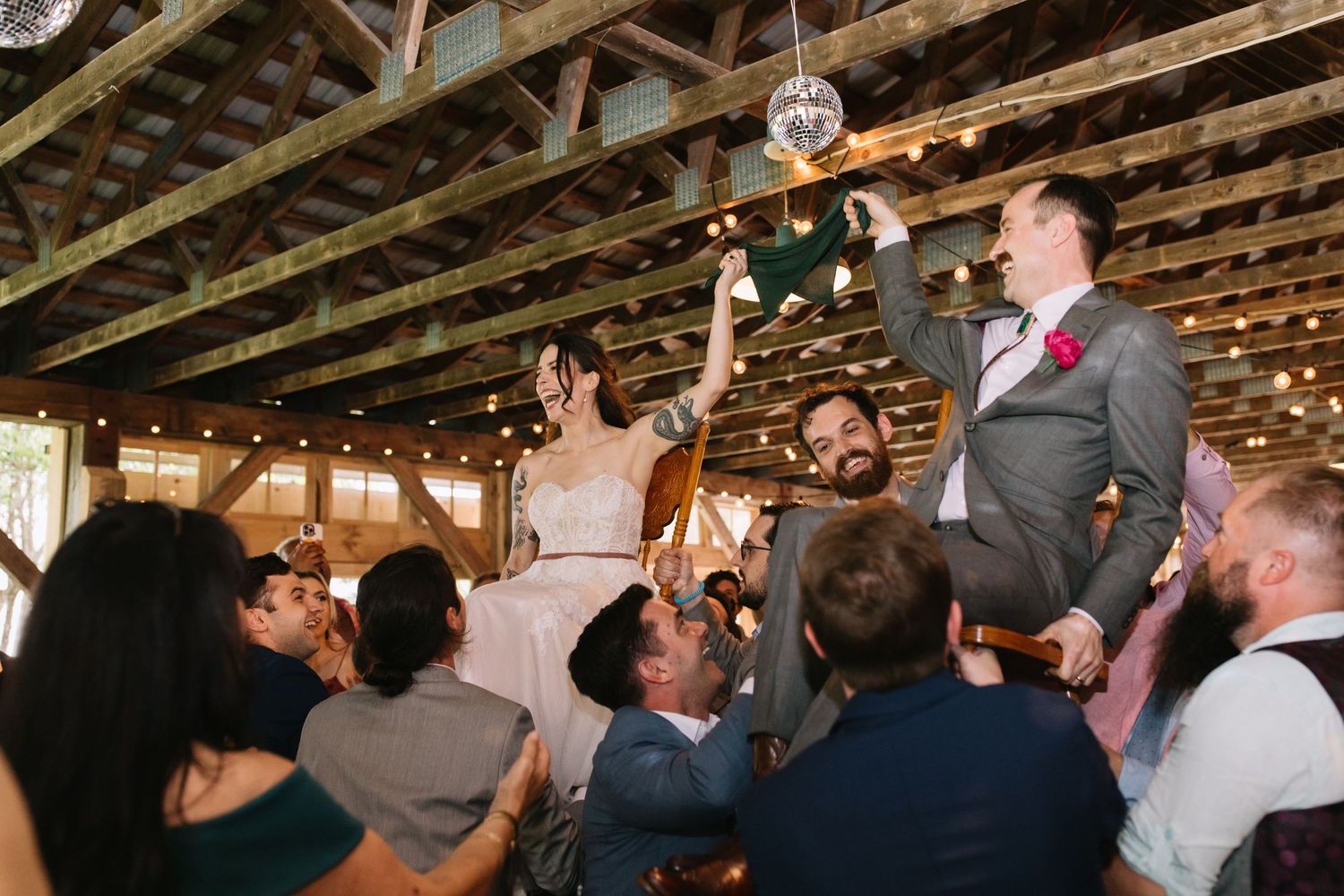 A group of wedding guests dance and celebrate together under wooden beams decorated with fairy lights at a reception.
