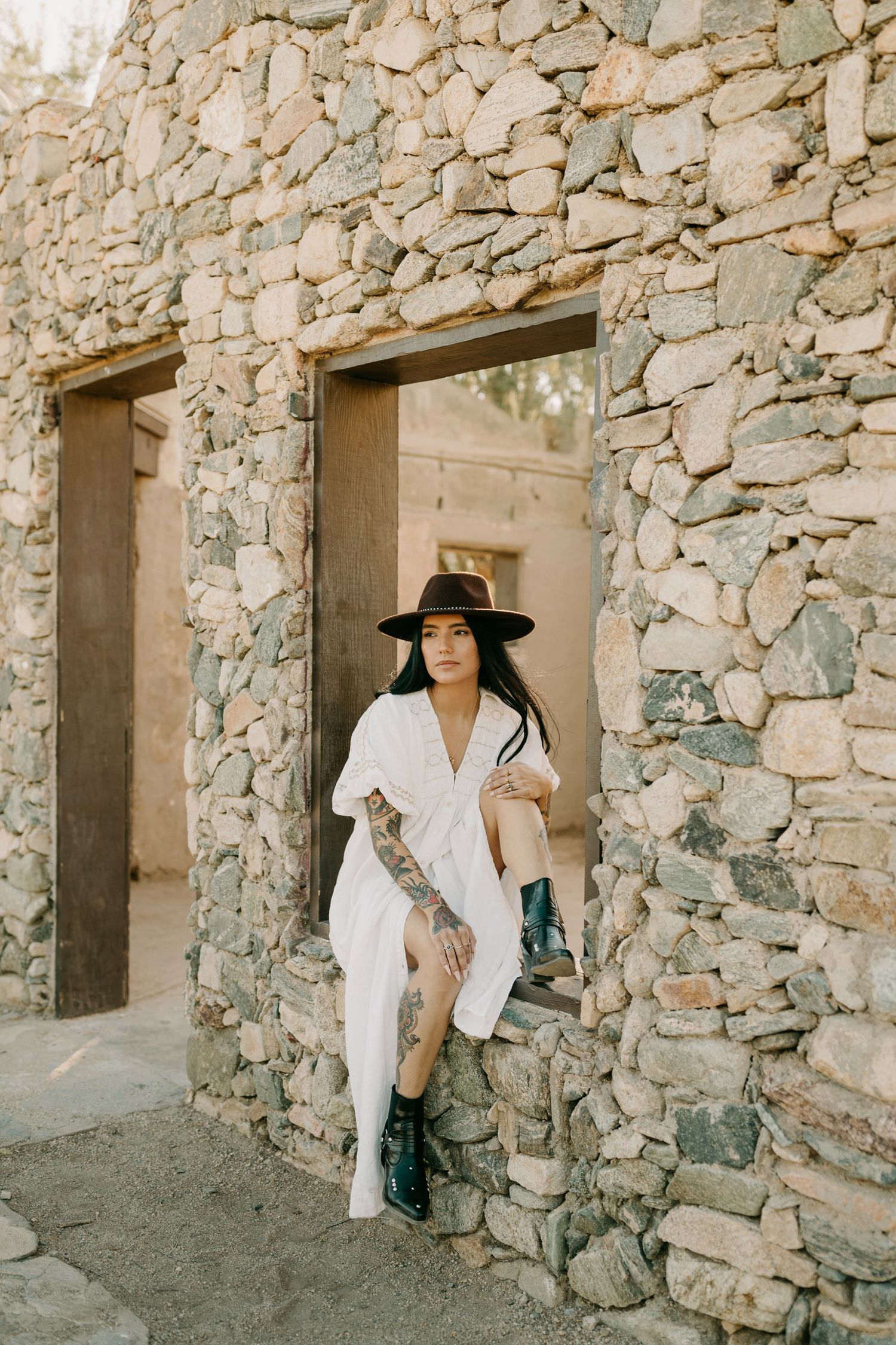 Person in white clothing and wide-brimmed hat poses in stone doorways of rustic building.