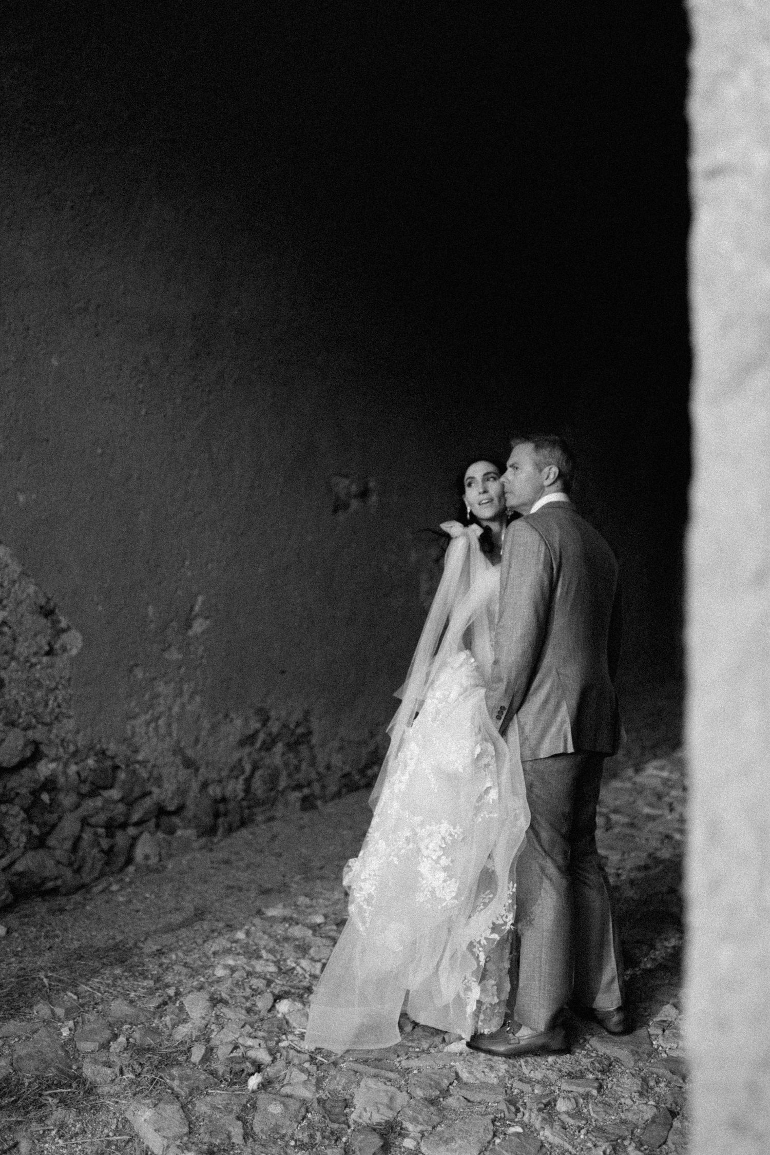 A black and white wedding photo of a couple in formalwear embracing against a textured dark wall.
