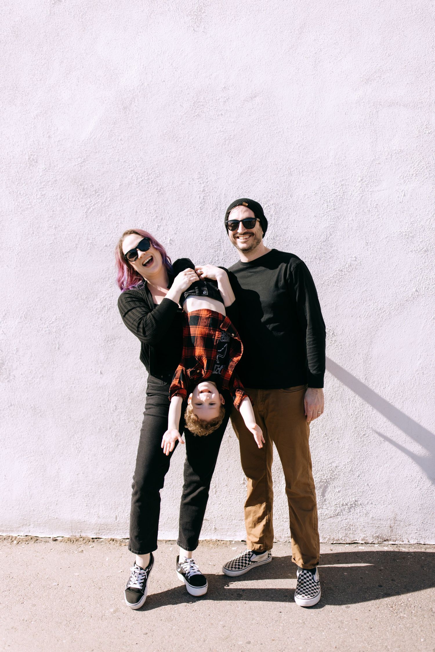 Two people wearing sunglasses and casual clothing pose playfully against a white wall in a series of minimalist photos.