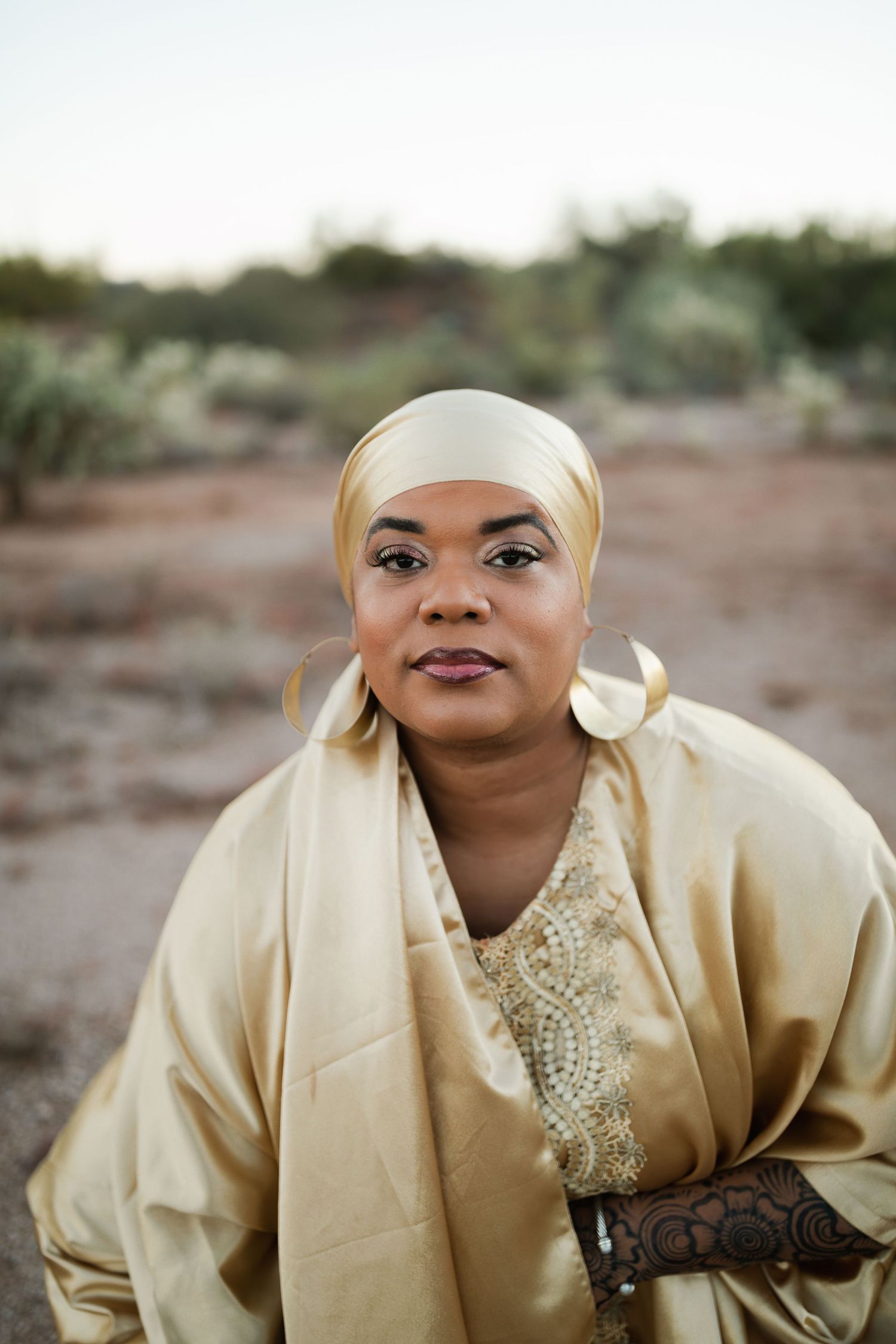 Portrait of someone in a cream colored headwrap and matching wrap dress with lace details in an outdoor desert setting.