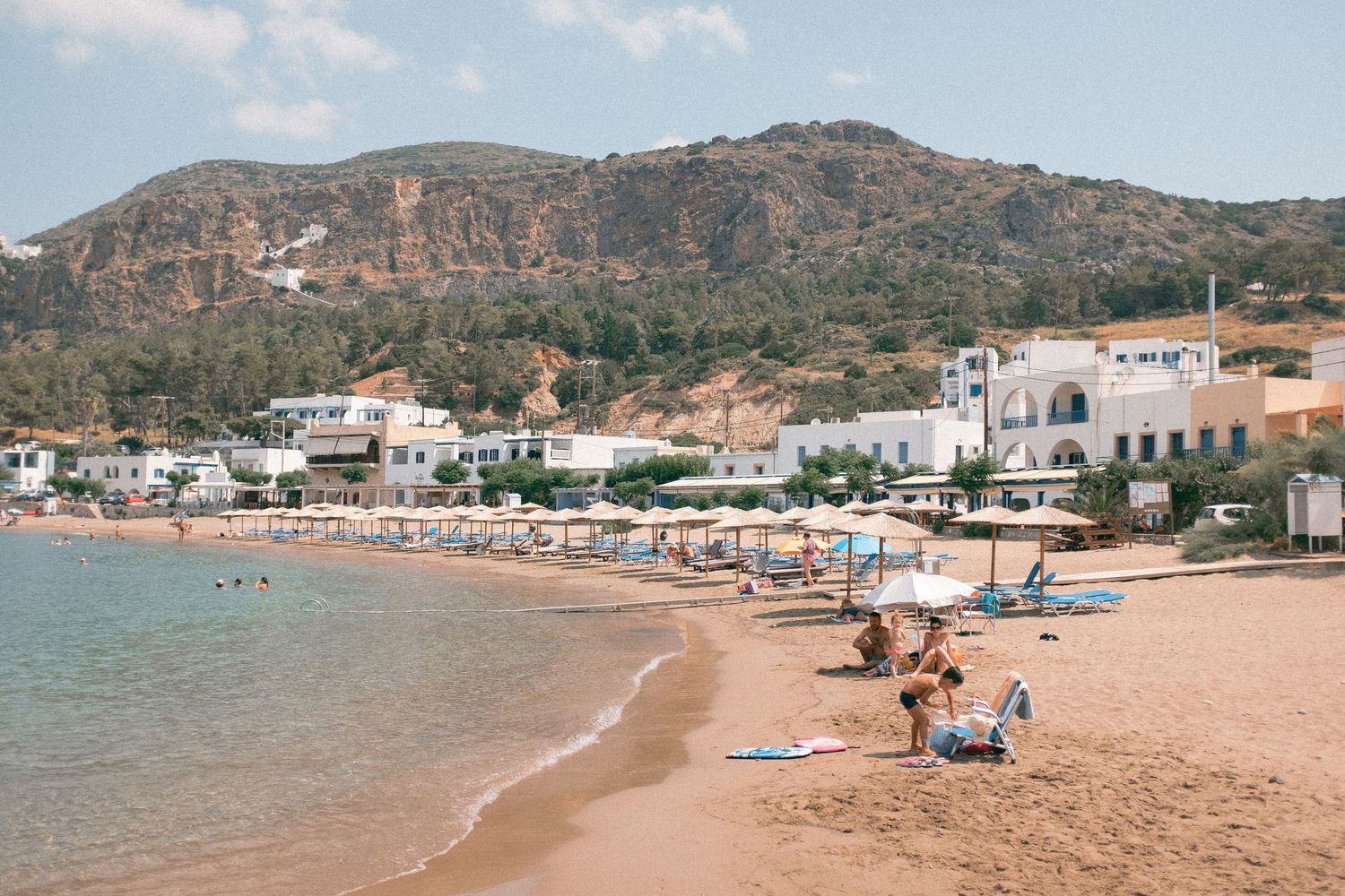 Sandy beach with umbrellas and loungers along Mediterranean coastline with white buildings and rocky hills in background.