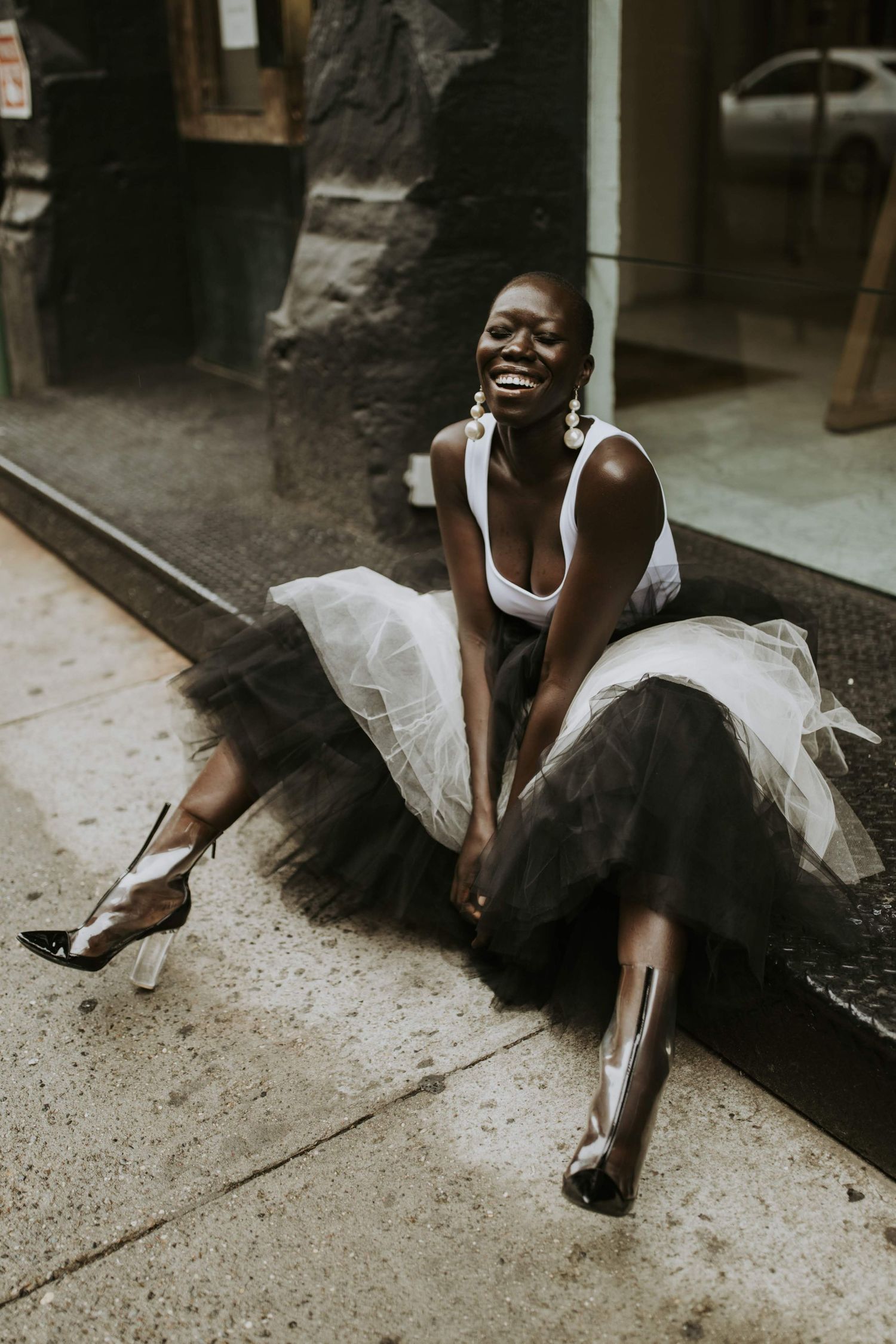 A dancer in a white top and tulle skirt sits on a sidewalk curb while laughing joyfully in an urban setting.