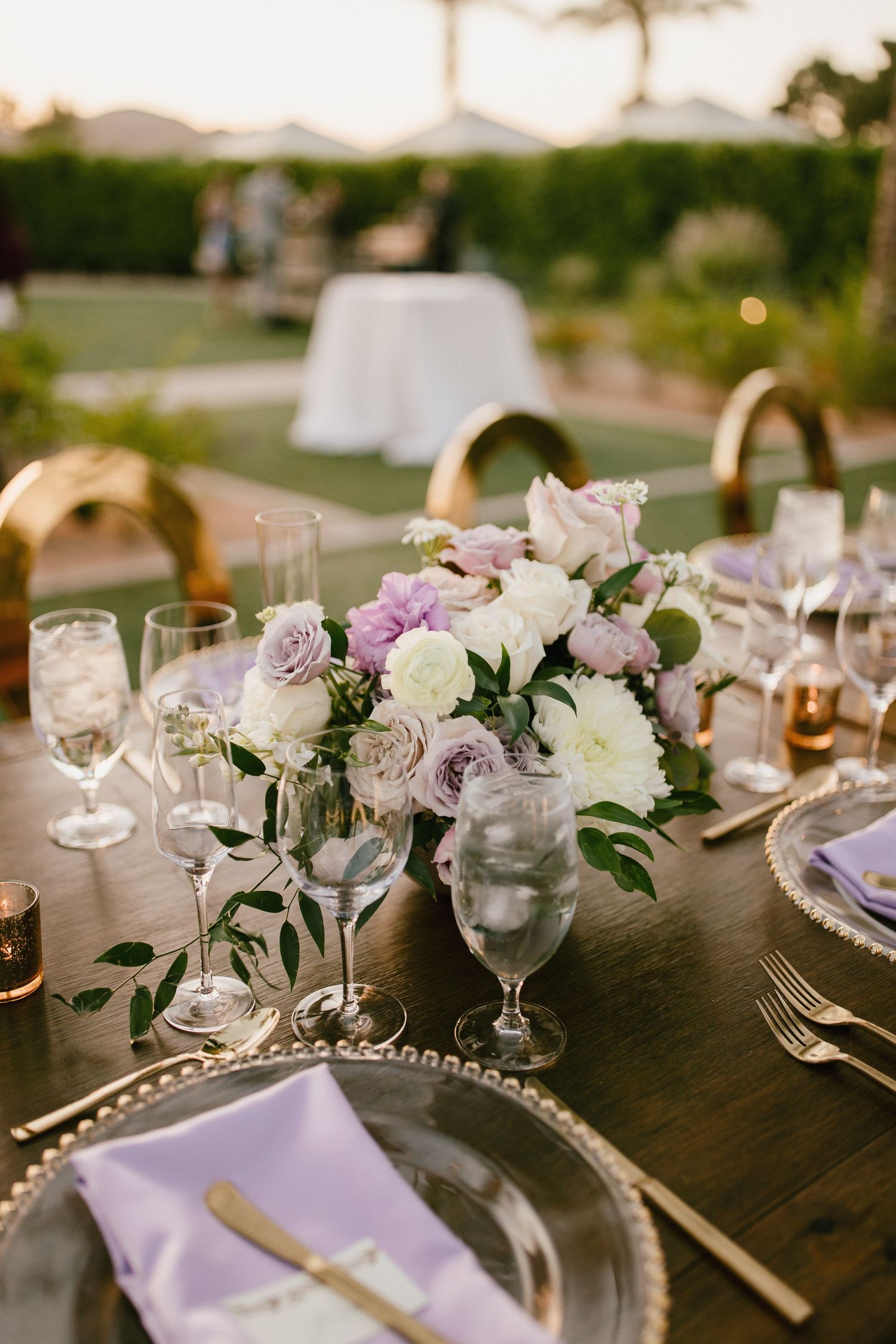 Elegant table setting with floral arrangements in purple and white colors, glassware, and silverware at dusk.