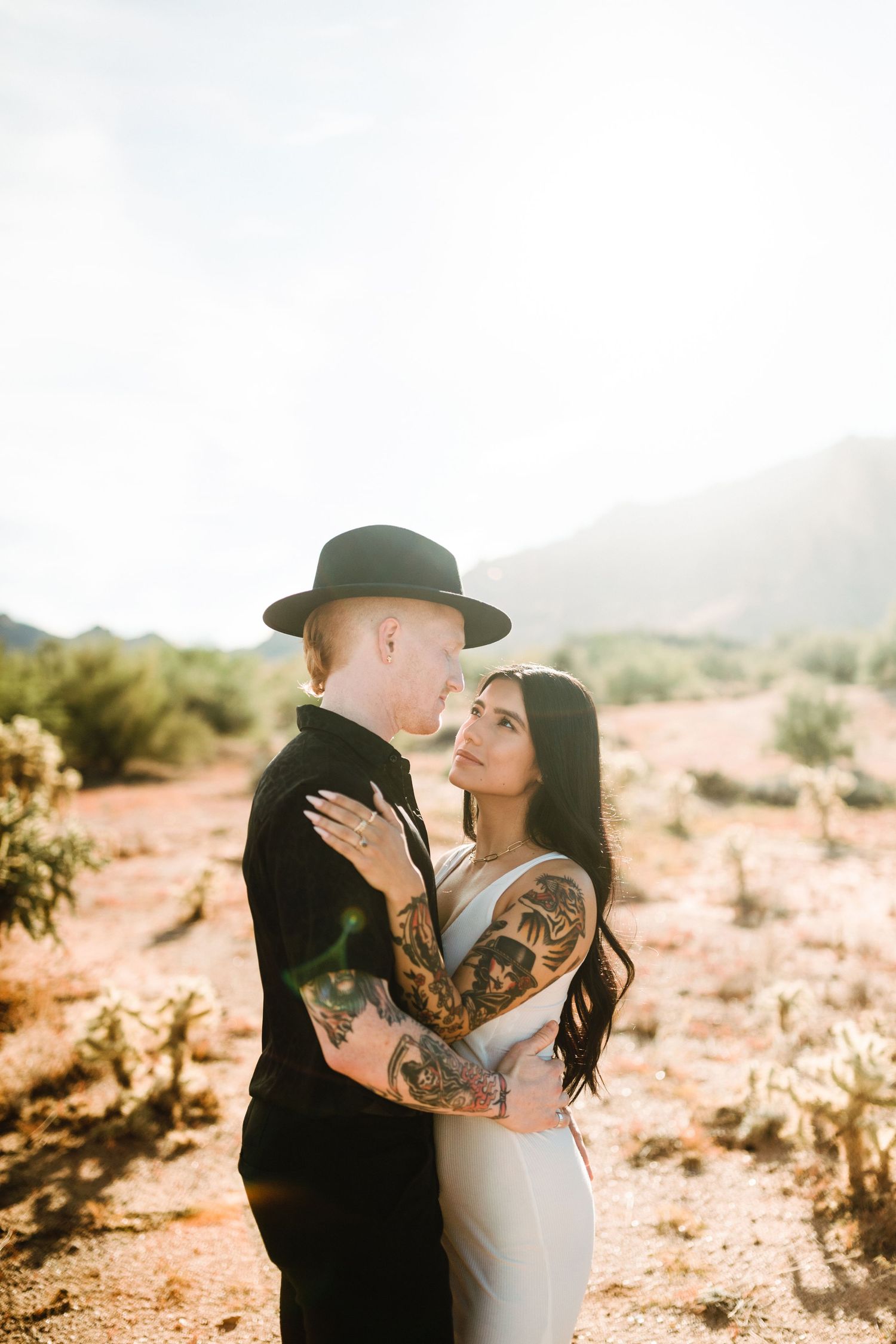 Desert engagement photo of a couple in black and white outfits embracing with visible tattoos on sunny day.