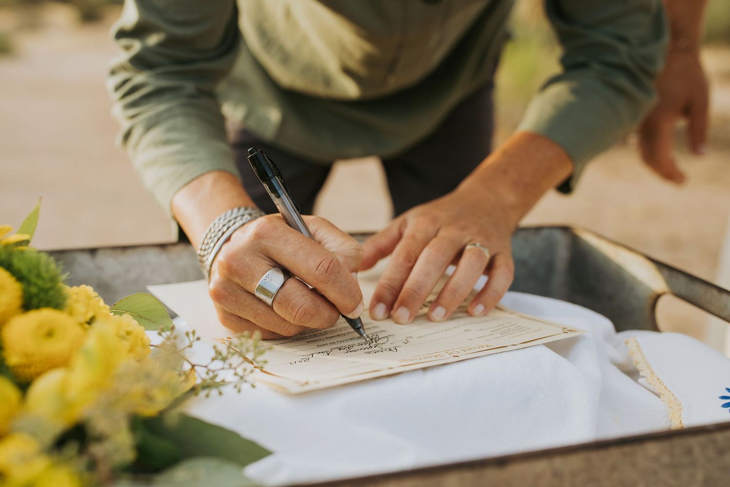 Someone wearing a sage green top writes with a pen on paper while yellow flowers sit nearby on the table.