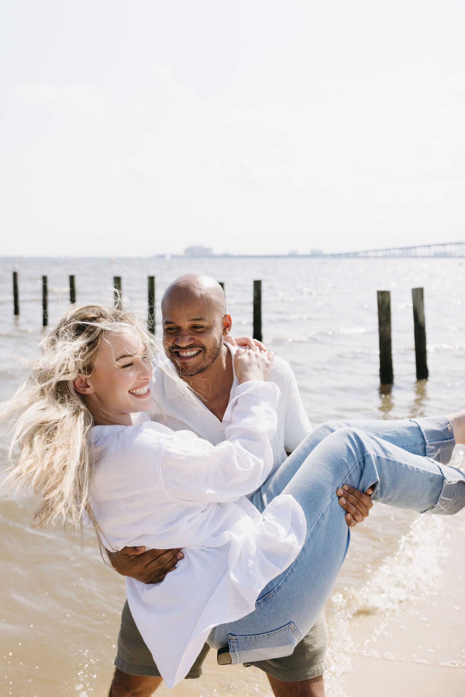 Happy couple laughing and embracing on sunny beach with wooden posts and water in background.