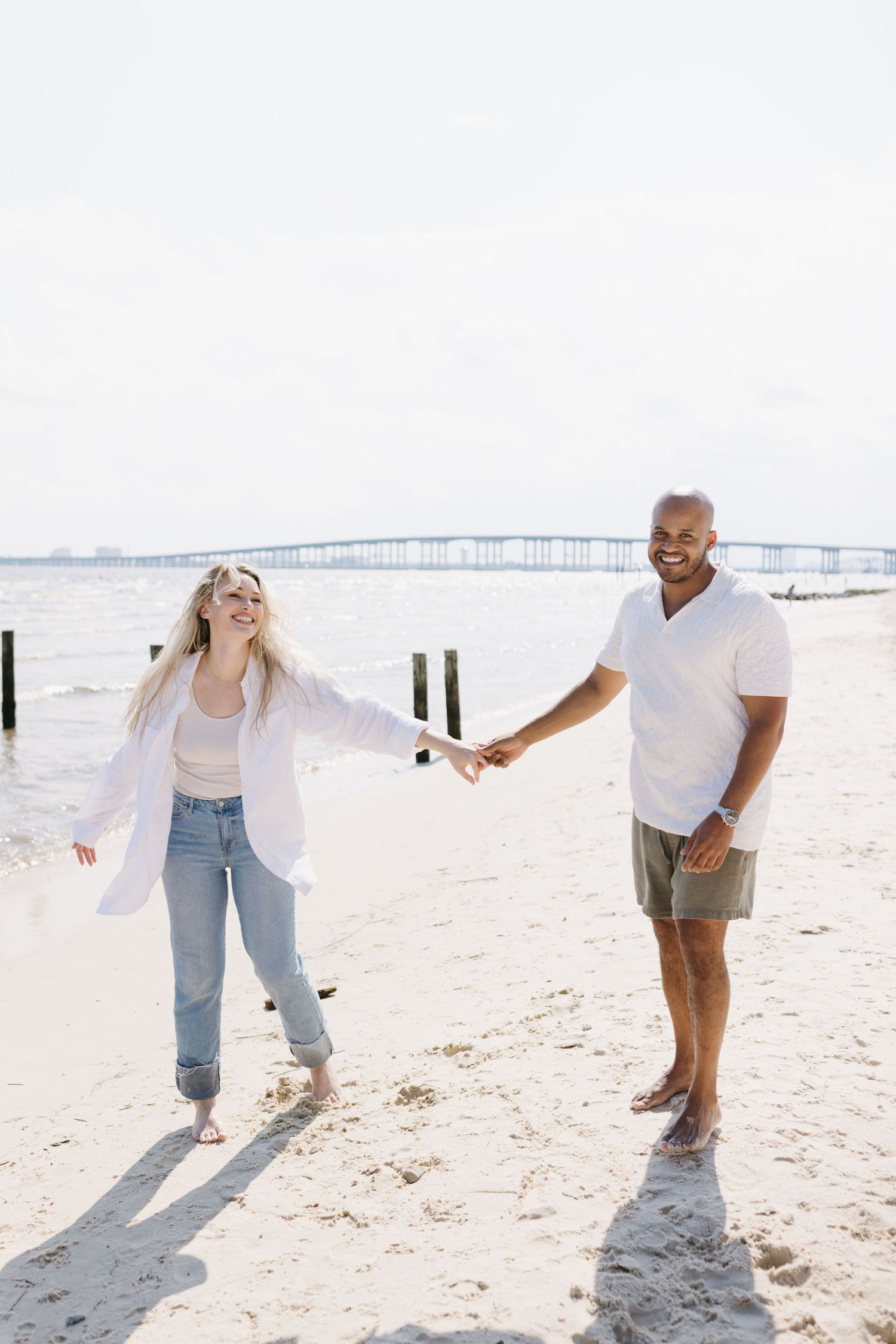 A couple holds hands while enjoying a sunny day at the beach with a bridge visible in the background.
