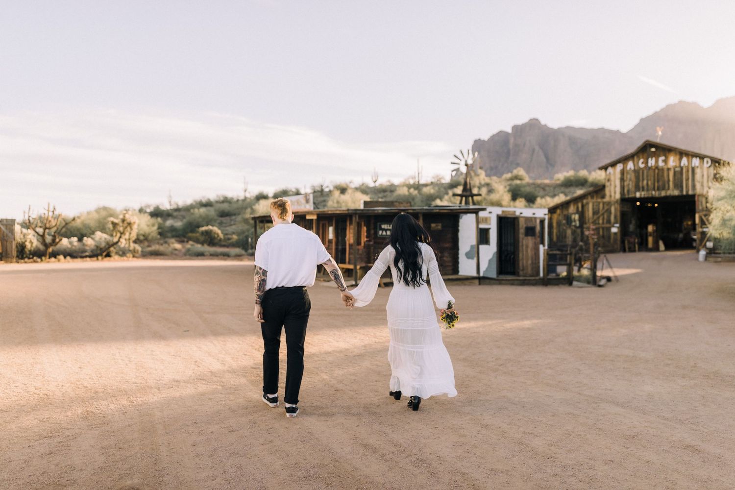 Bride and groom walking hand in hand across a desert landscape with mountains in the background at sunset.