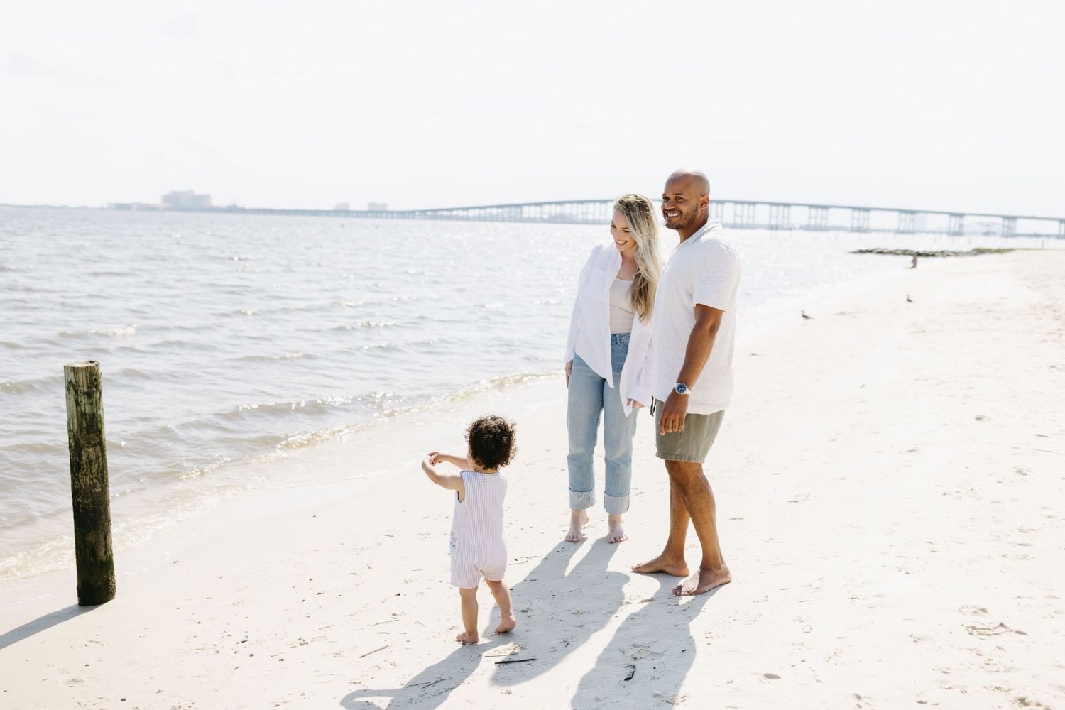 A small family stands together on a sunny white sand beach with calm ocean waters in the background.