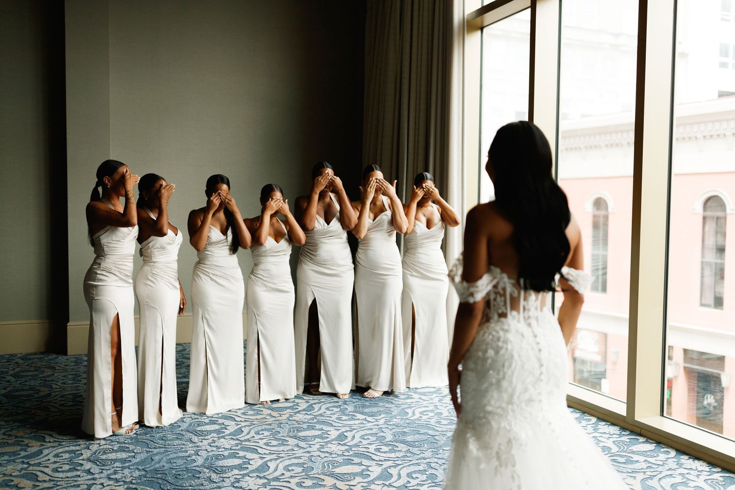 Bridesmaids in matching white gowns turn their backs while bride in lace dress watches them by large window.