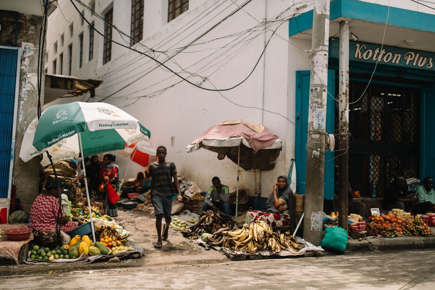 A panoramic view of a street market with vendors selling fresh produce and goods beneath colorful awnings.