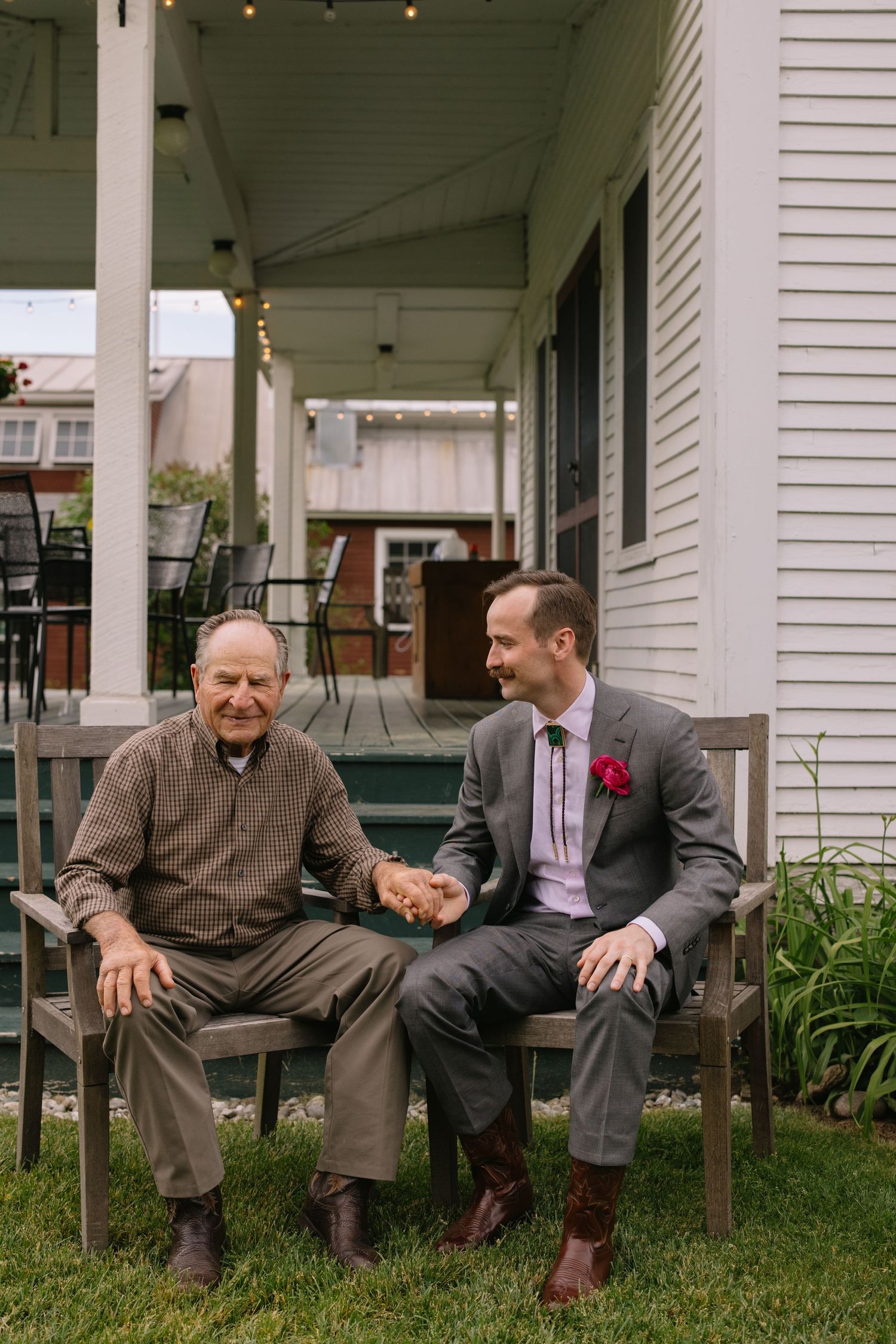 A senior and younger man in a suit sit together on a wooden bench on a porch with white siding.