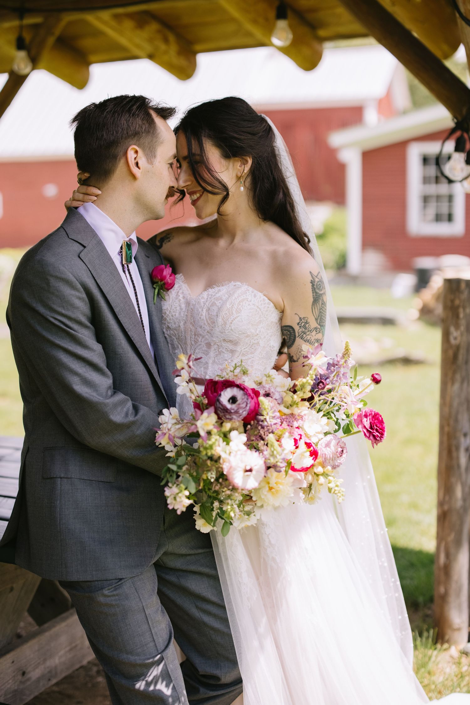 Newlyweds share intimate moment under wooden shelter with red barn in background and colorful floral bouquet.