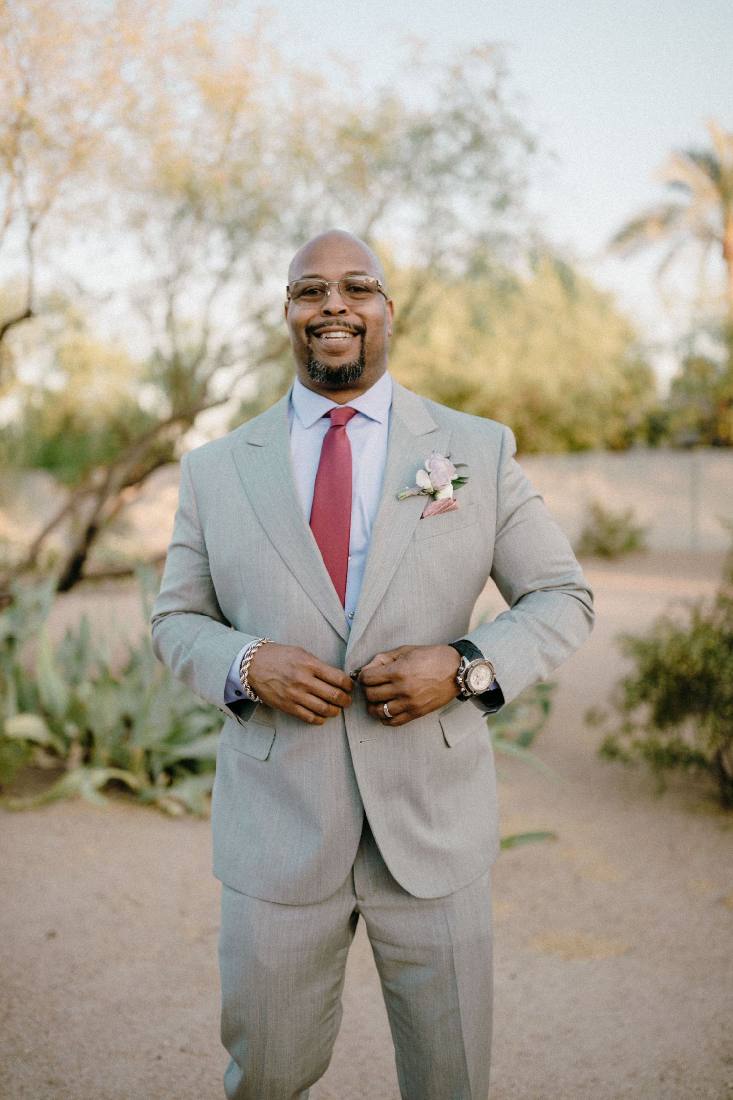 A wedding party member smiles while adjusting his light gray suit jacket and red tie outdoors at a desert venue.