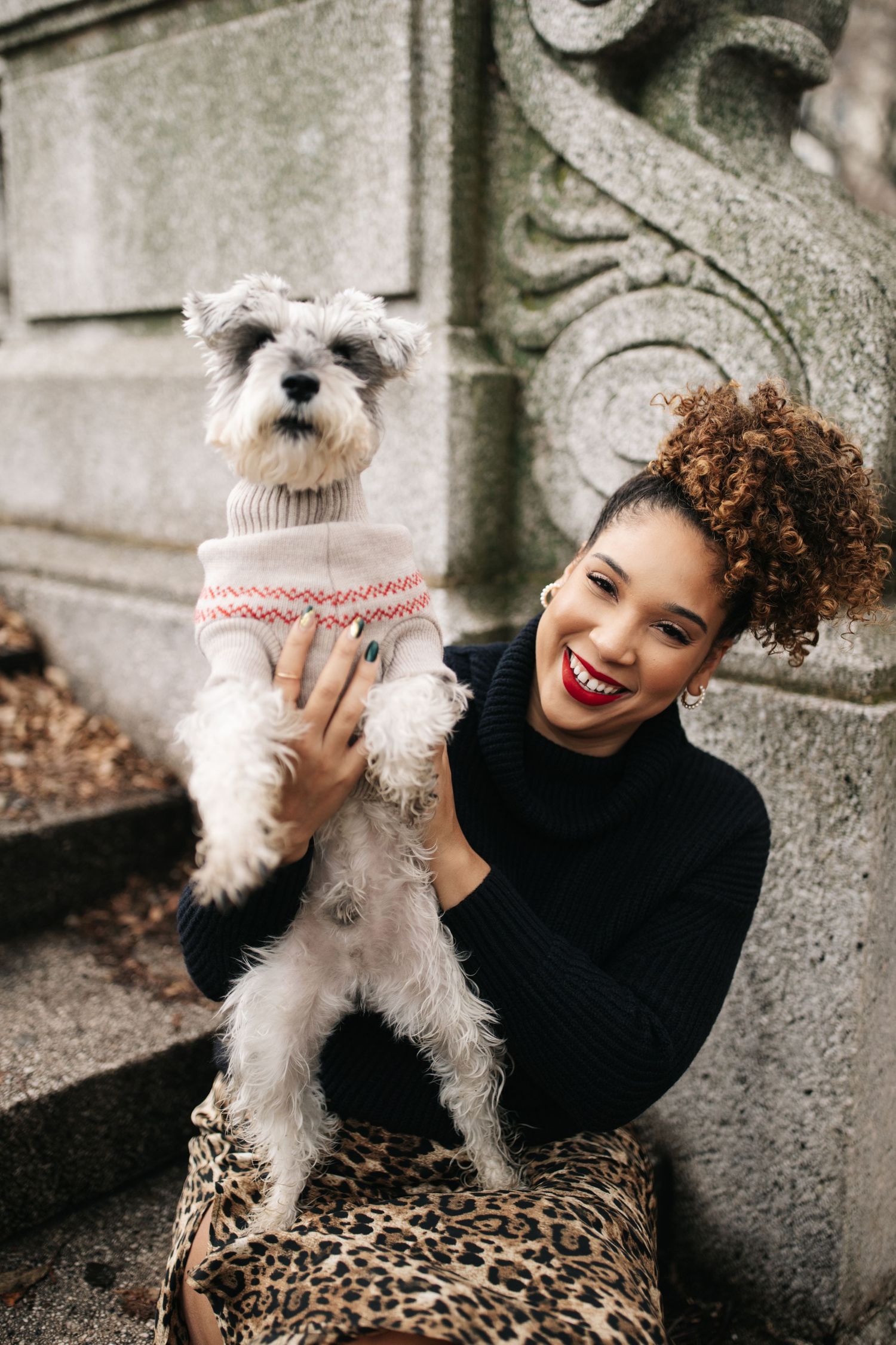 A smiling person in black sweater poses with small white Maltese dog wearing a patterned sweater for a winter photoshoot.