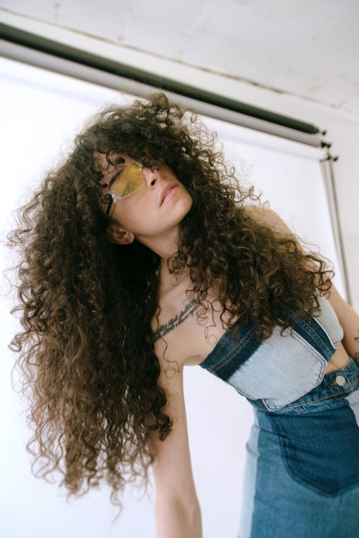 Fashion portrait featuring dramatic curly hair and denim overalls against a white wall interior.