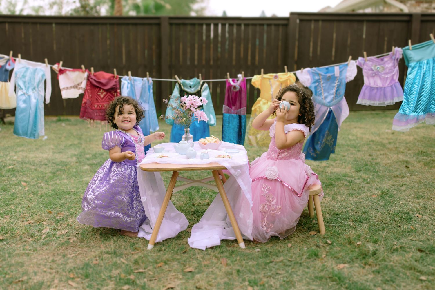 Two dolls wearing princess dresses having tea at a small table with clothes hanging on a line in the background.